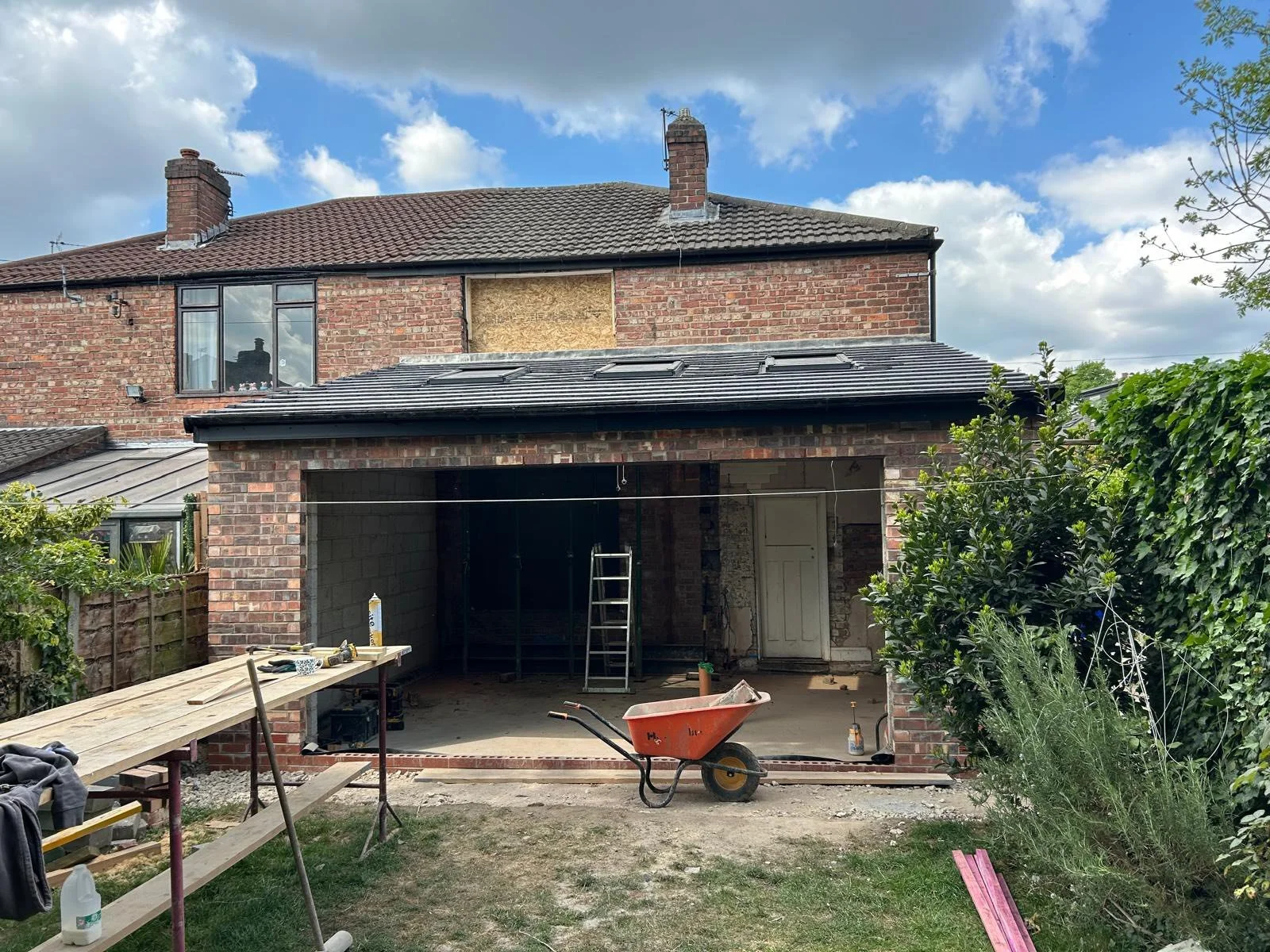 Rear view of a two-story brick house undergoing renovation with an extension being added. The extension has a black tiled roof, and the back wall is open for construction. Scaffolding, a ladder, wheelbarrow, construction tools, and materials are visible. The sky is partly cloudy with trees and bushes around.