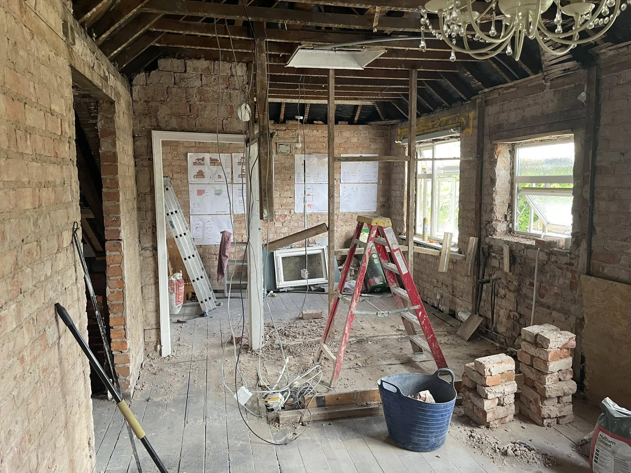 Room under renovation with exposed brick walls, construction tools, a ladder, scattered bricks, and electrical wiring hanging from the ceiling.