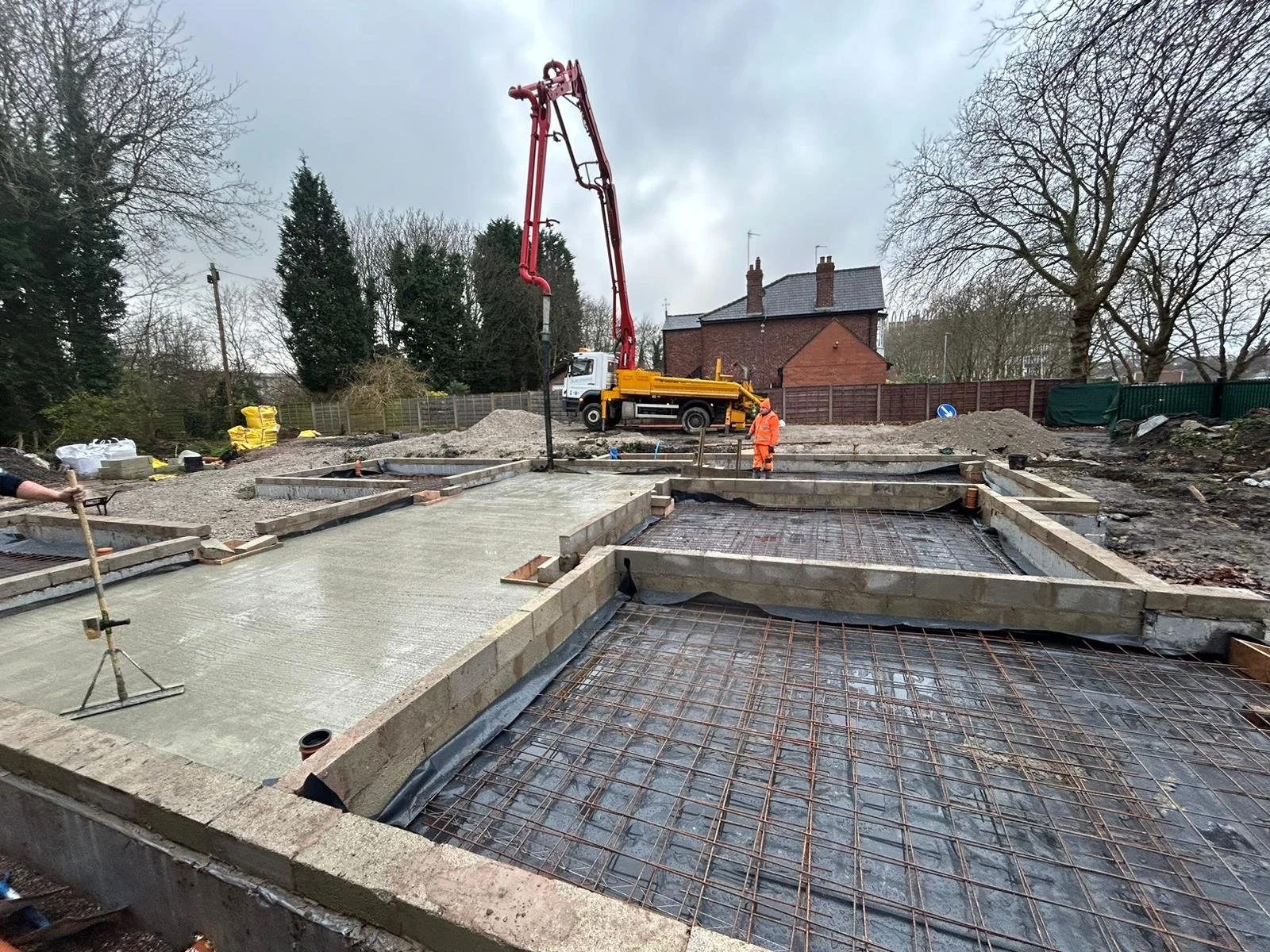 Construction site with concrete pouring and reinforcement for building foundations, worker in orange safety gear, concrete pump truck in background, and partly built concrete slabs with rebar.