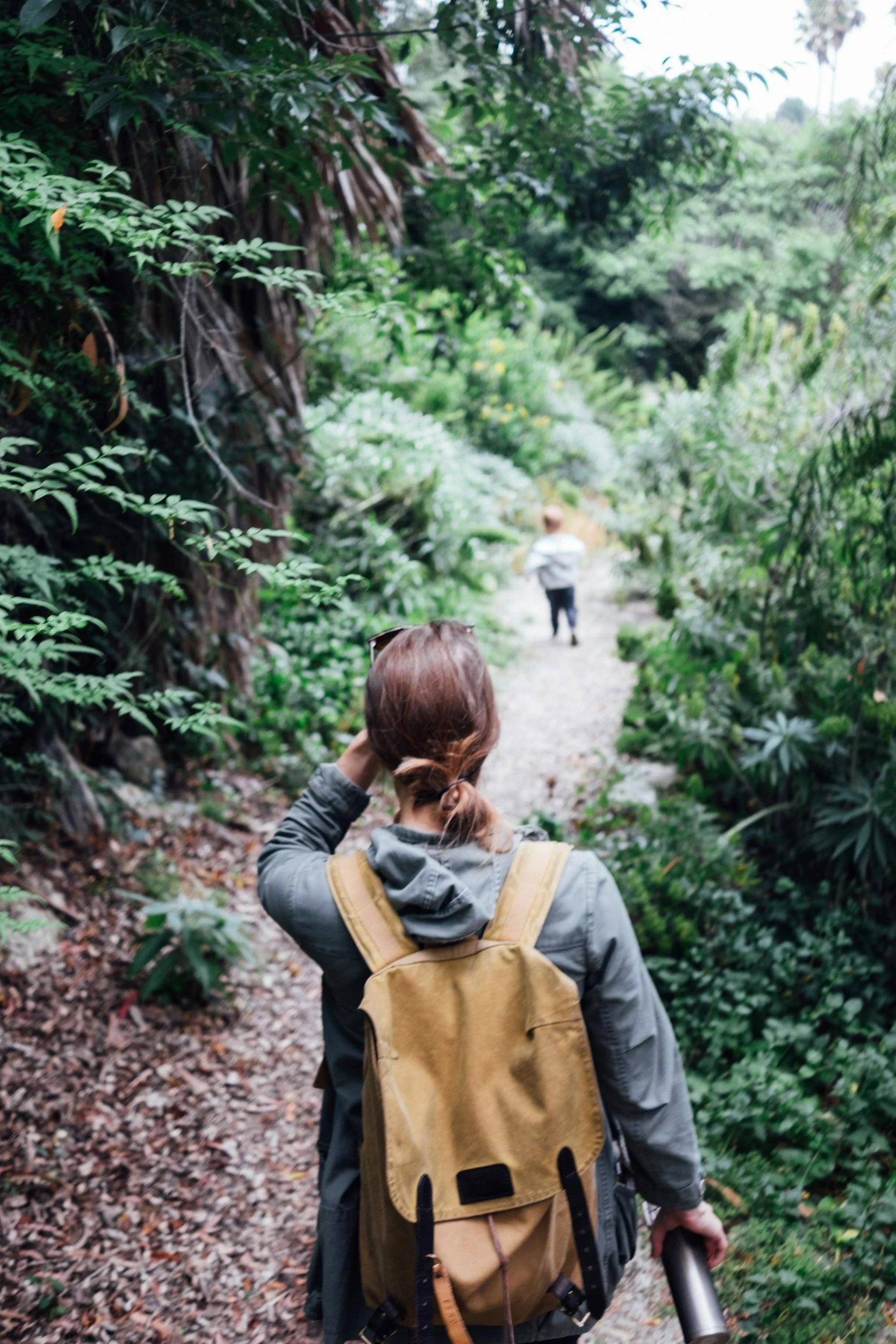 A woman with a tan backpack walking on a forest trail, holding a thermos, and looking at a person ahead of her in a lush green forest.