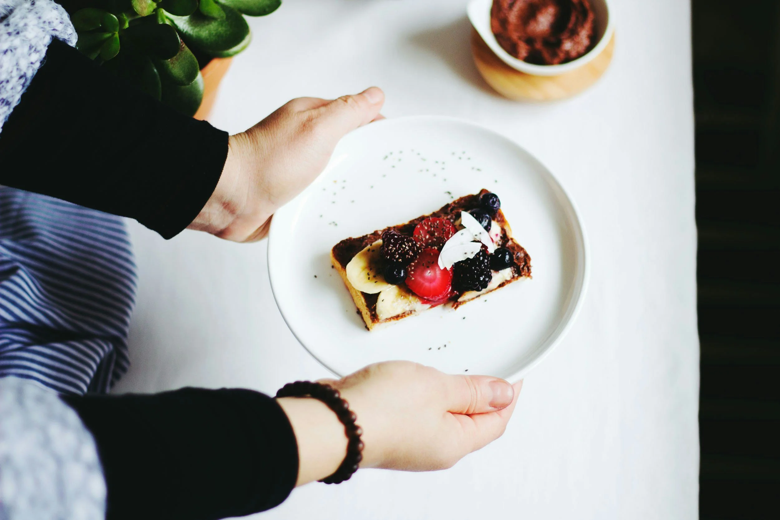 Person holding a white plate with a slice of dessert topped with berries and fruit, on a white table with a small bowl of more berries and green plants nearby.