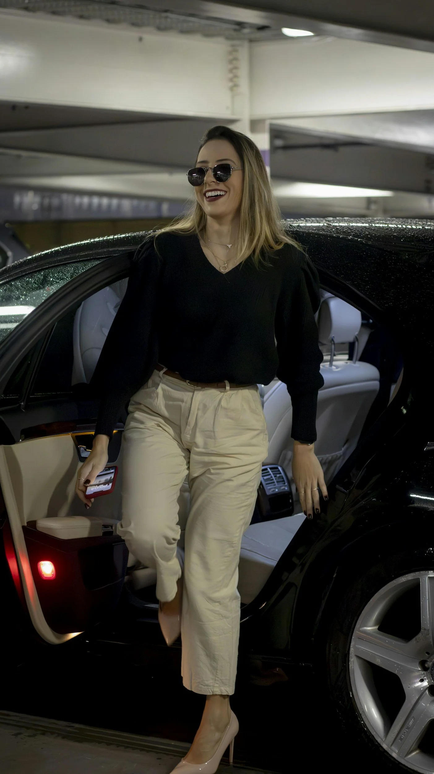 A woman with blonde hair, wearing sunglasses, a black top, light beige pants, and high heels, standing next to a black luxury car in an indoor parking garage.