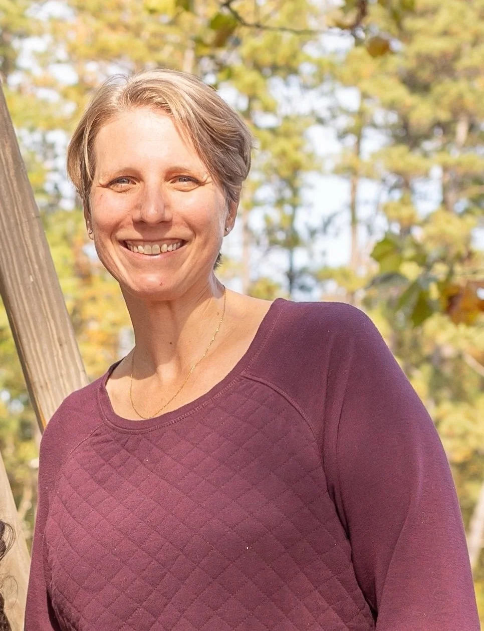 A woman with short blonde hair smiling outdoors in a park with trees and autumn foliage in the background.
