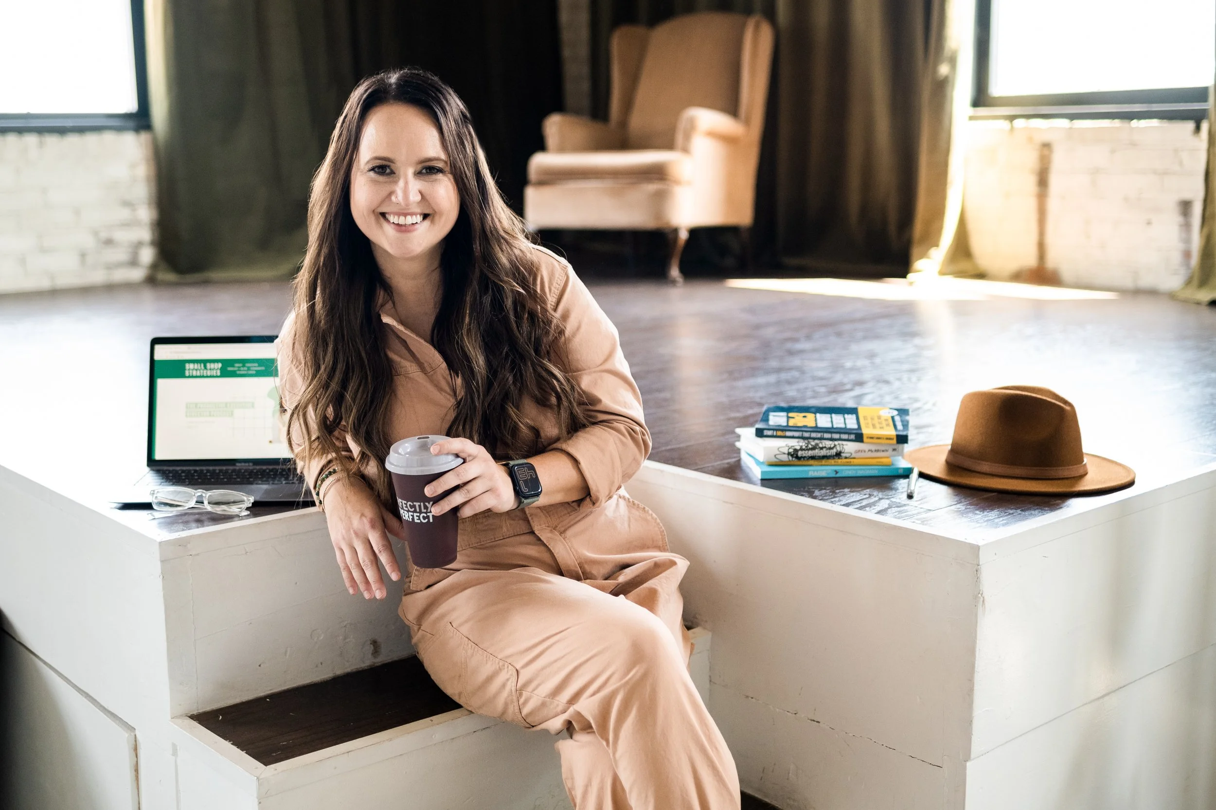 Rachel Bearbower of Nonprofit Automation Agency sitting on a white ledge, holding a coffee cup, with a laptop, books, and a hat nearby in an airy room with large windows.