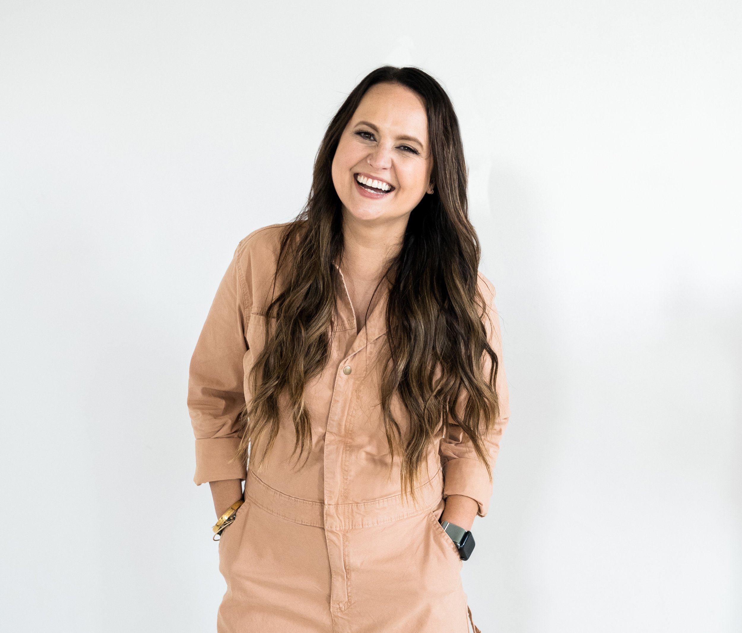 Woman with long brown hair smiling, wearing a tan jumpsuit with hands in pockets, standing against a plain white background.