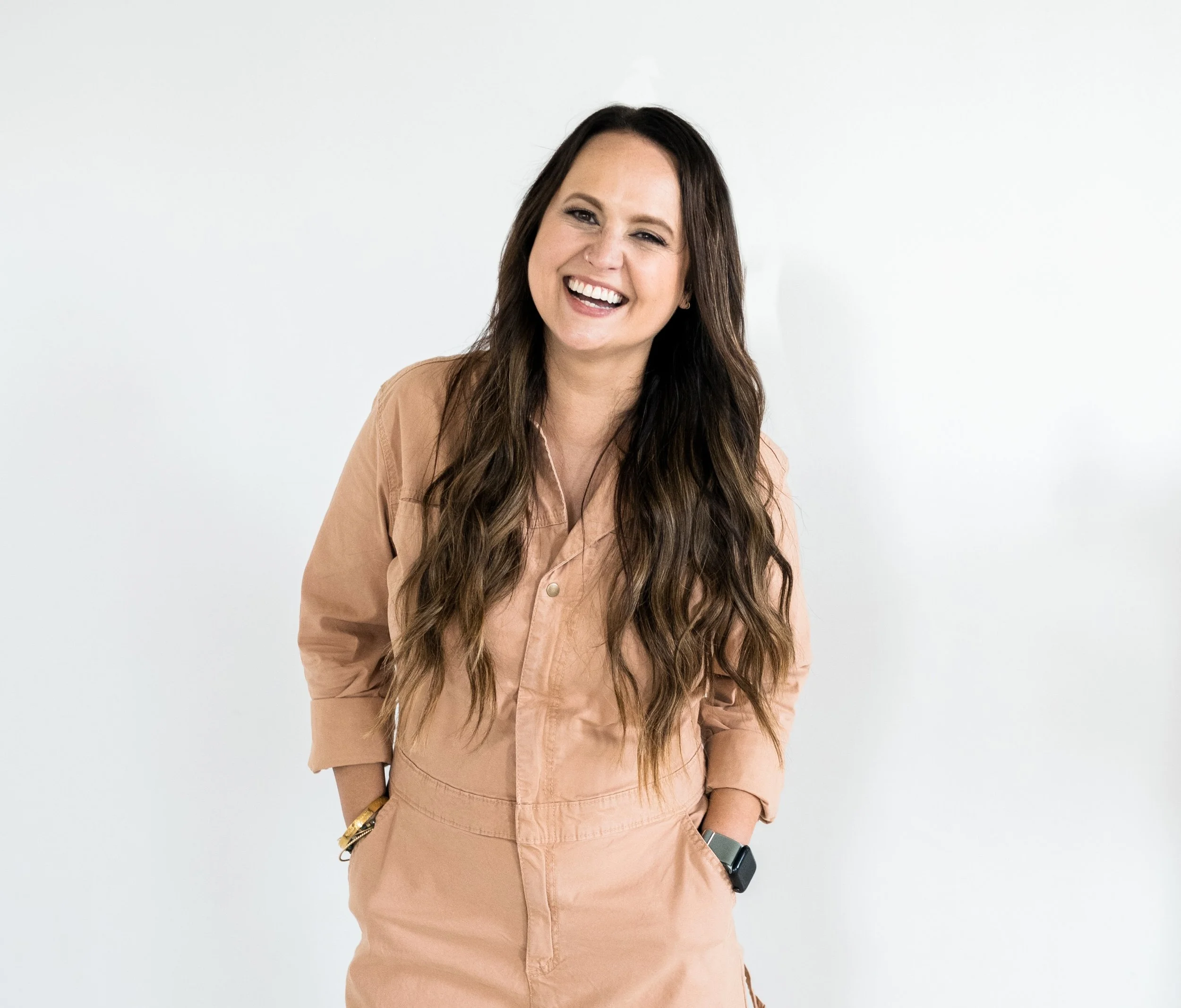 A woman with long wavy brown hair is smiling and standing against a plain white background. She is wearing a beige jumpsuit with the sleeves rolled up and has her hands in her pockets.