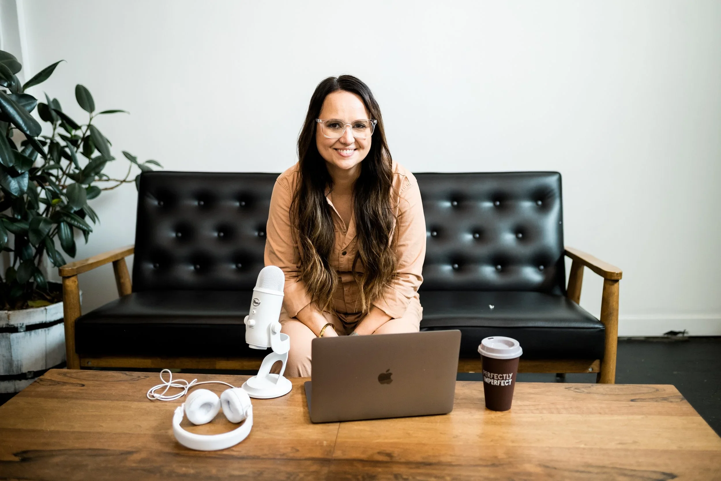 Woman sitting on a black leather couch with a wooden frame, smiling, with a microphone, laptop, headphones, and a coffee cup on the wooden table in front of her, in an office or studio setting.