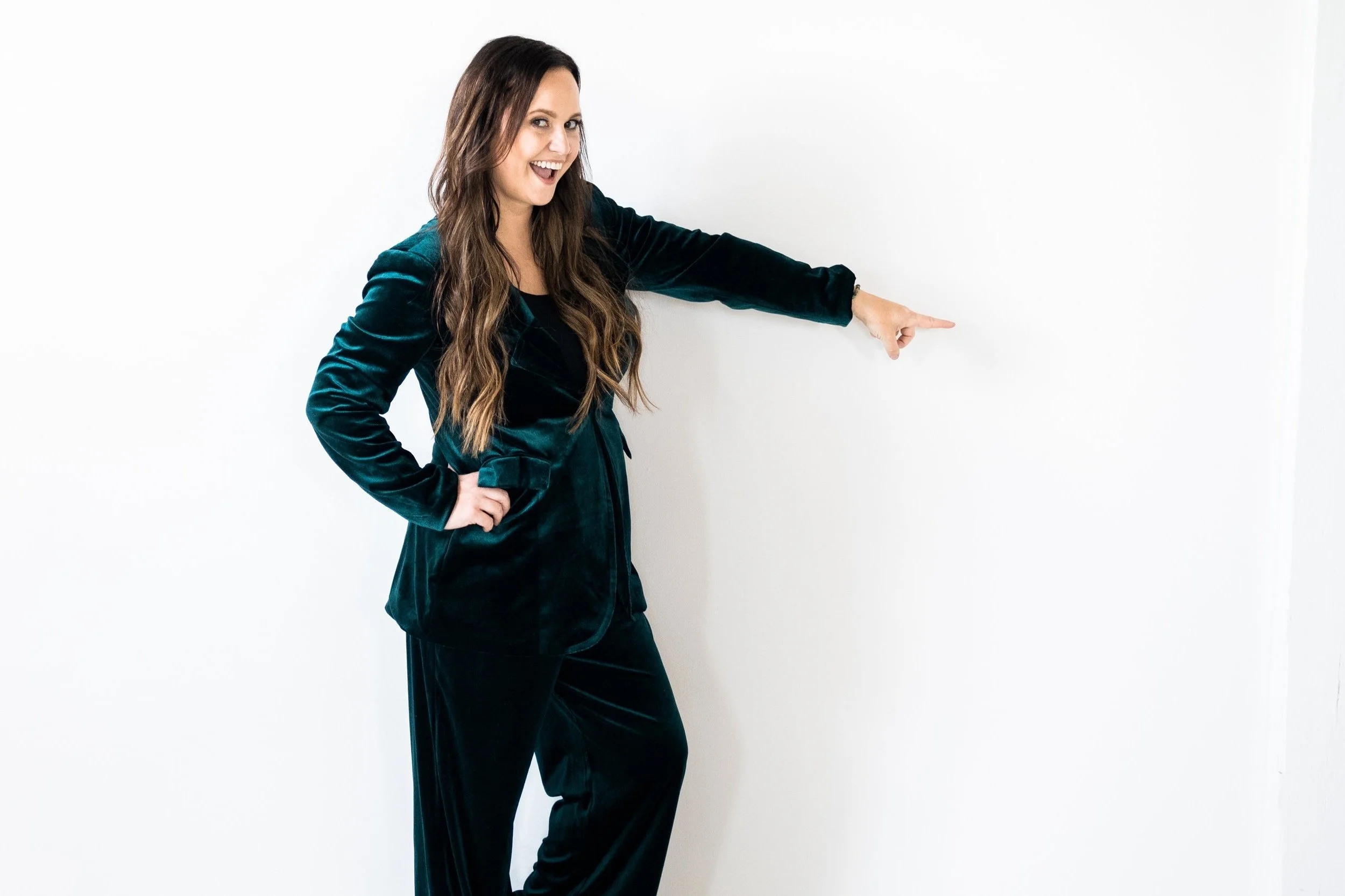 Rachel Bearbower of Nonprofit Automation Agency wearing a teal velvet blazer and black pants, smiling and pointing towards the right side of the image against a plain white background.