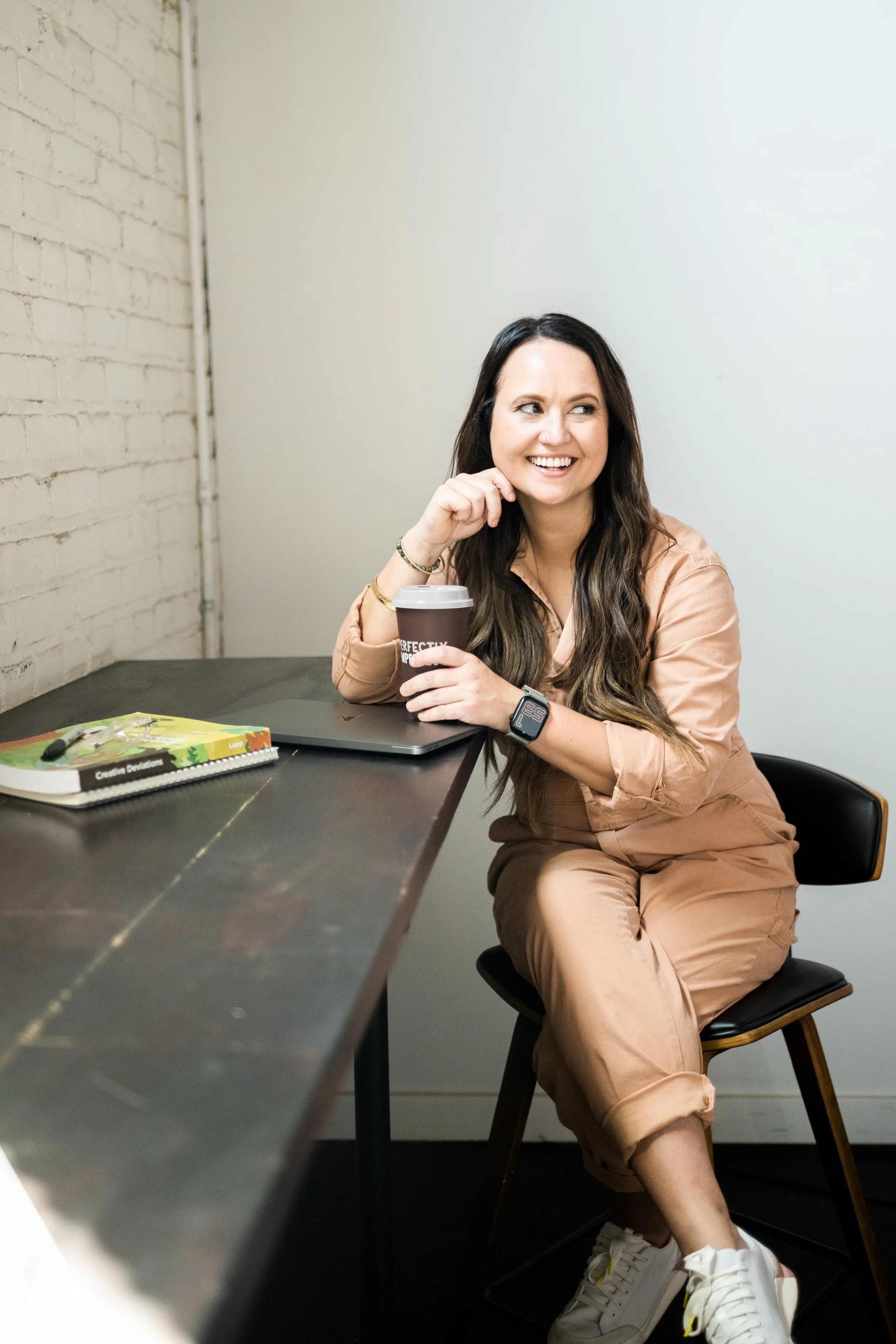 A woman with long dark hair sitting at a desk, holding a coffee cup and smiling.