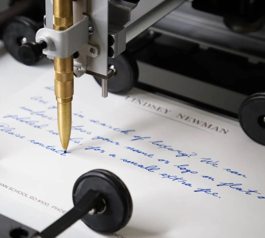 Close-up of a handwriting machine with a golden pen writing blue ink cursive text on white paper.