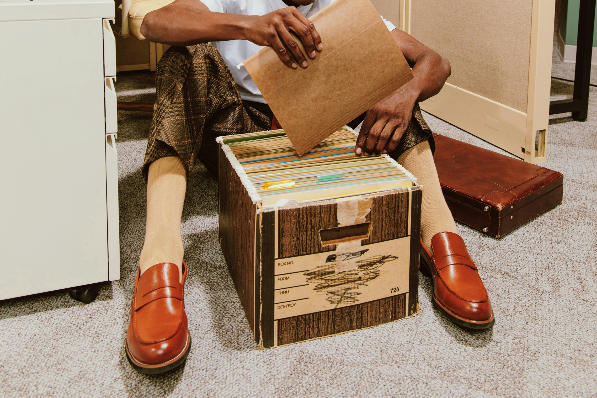 A person sitting on the floor, browsing through a large cardboard box filled with files or folders, surrounded by office furniture and briefcases.
