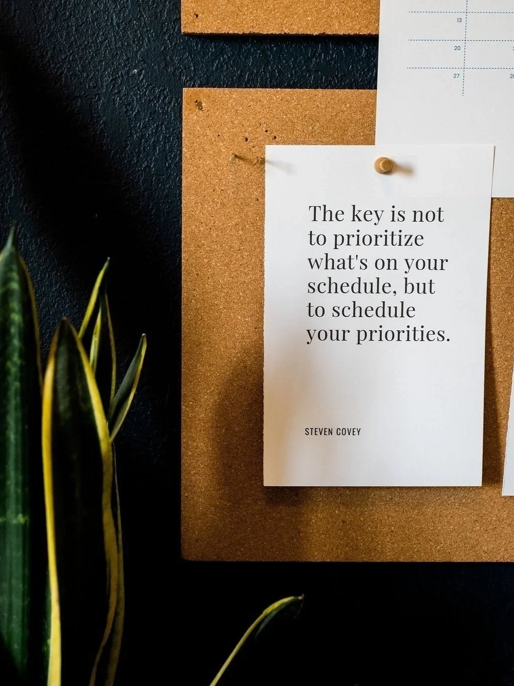 Quote on a cork board that says, "The key is not to prioritize what's on your schedule, but to schedule your priorities," by Steven Covey. Part of a calendar and a green plant are visible.