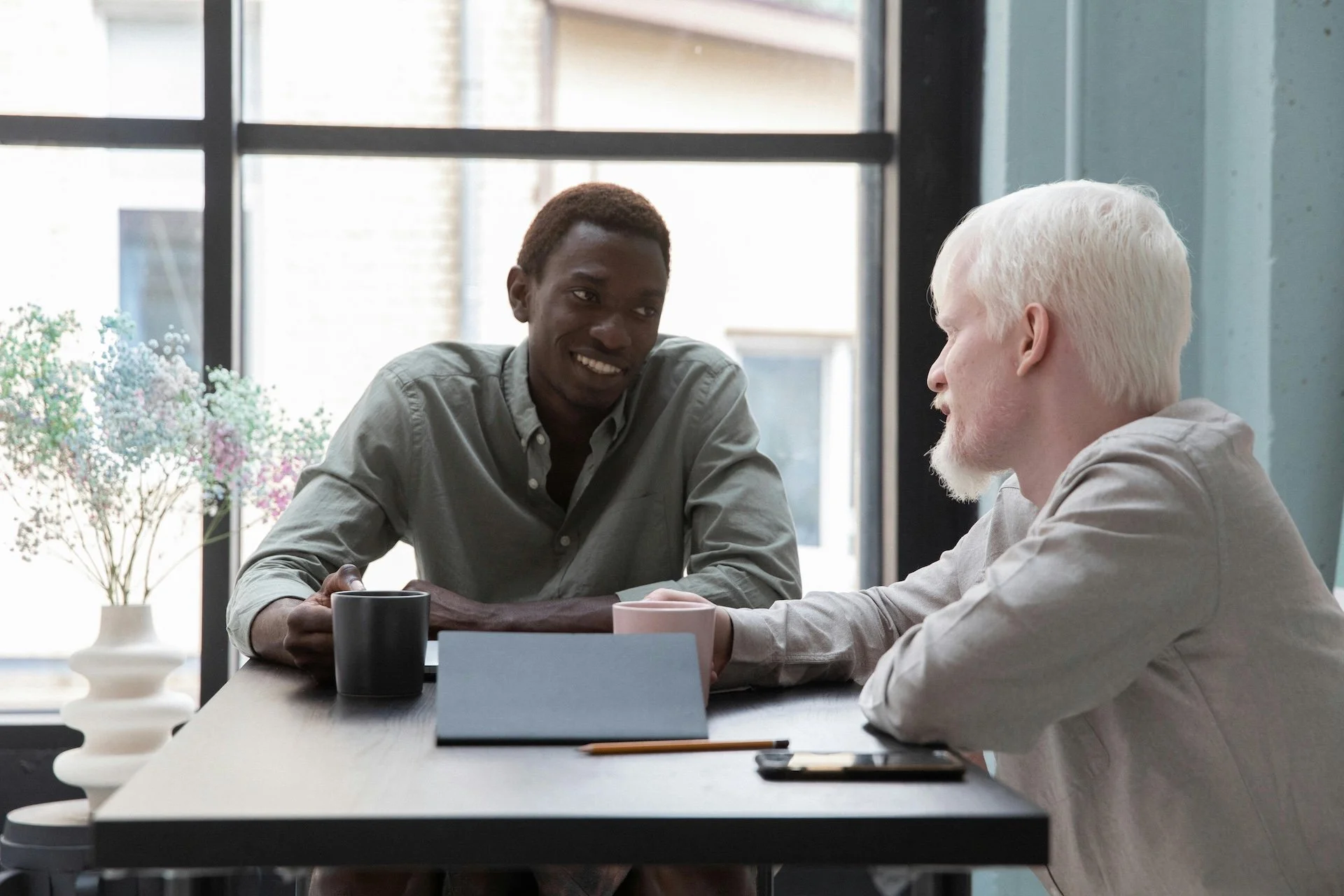 Two men sitting at a table engaged in conversation, with coffee mugs, a tablet, and a smartphone on the table in a well-lit room with large windows.