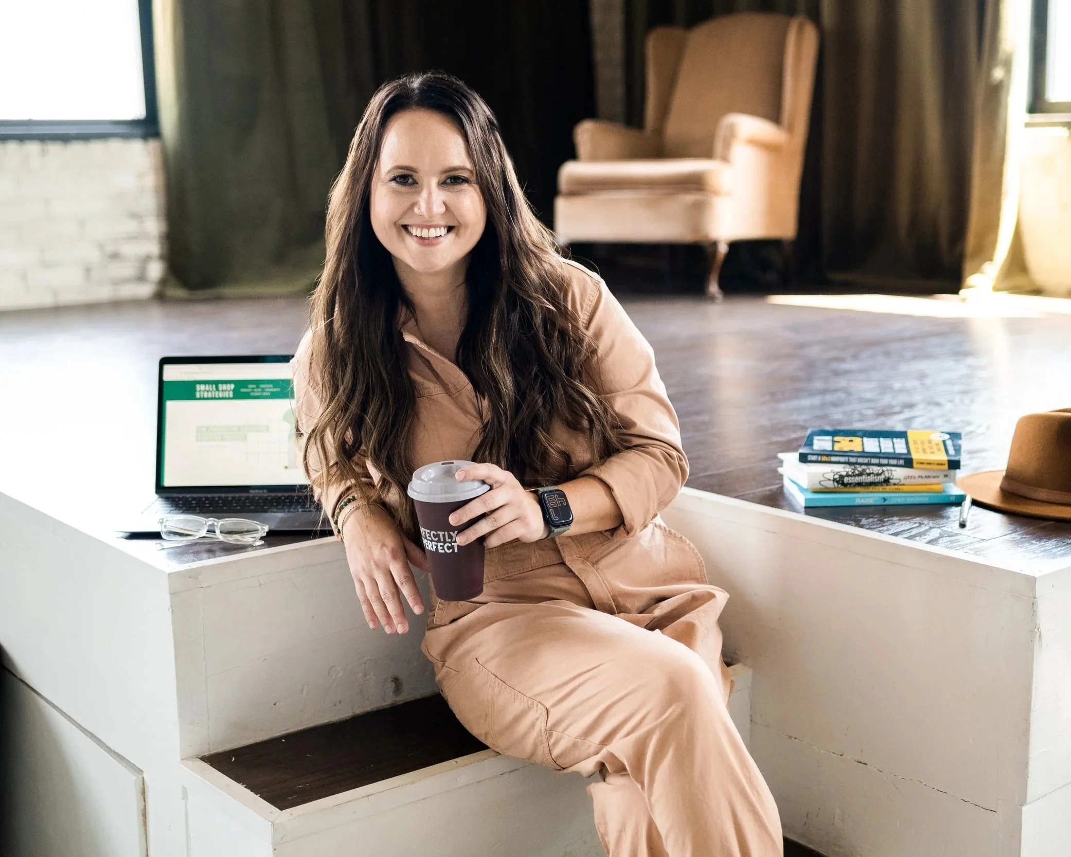 A woman with long brown hair, smiling, sitting on a white bench indoors. She is holding a paper coffee cup and wearing a tan jumpsuit and a smartwatch. There are books, a hat, and eyeglasses on the bench beside her, and a laptop open behind her.