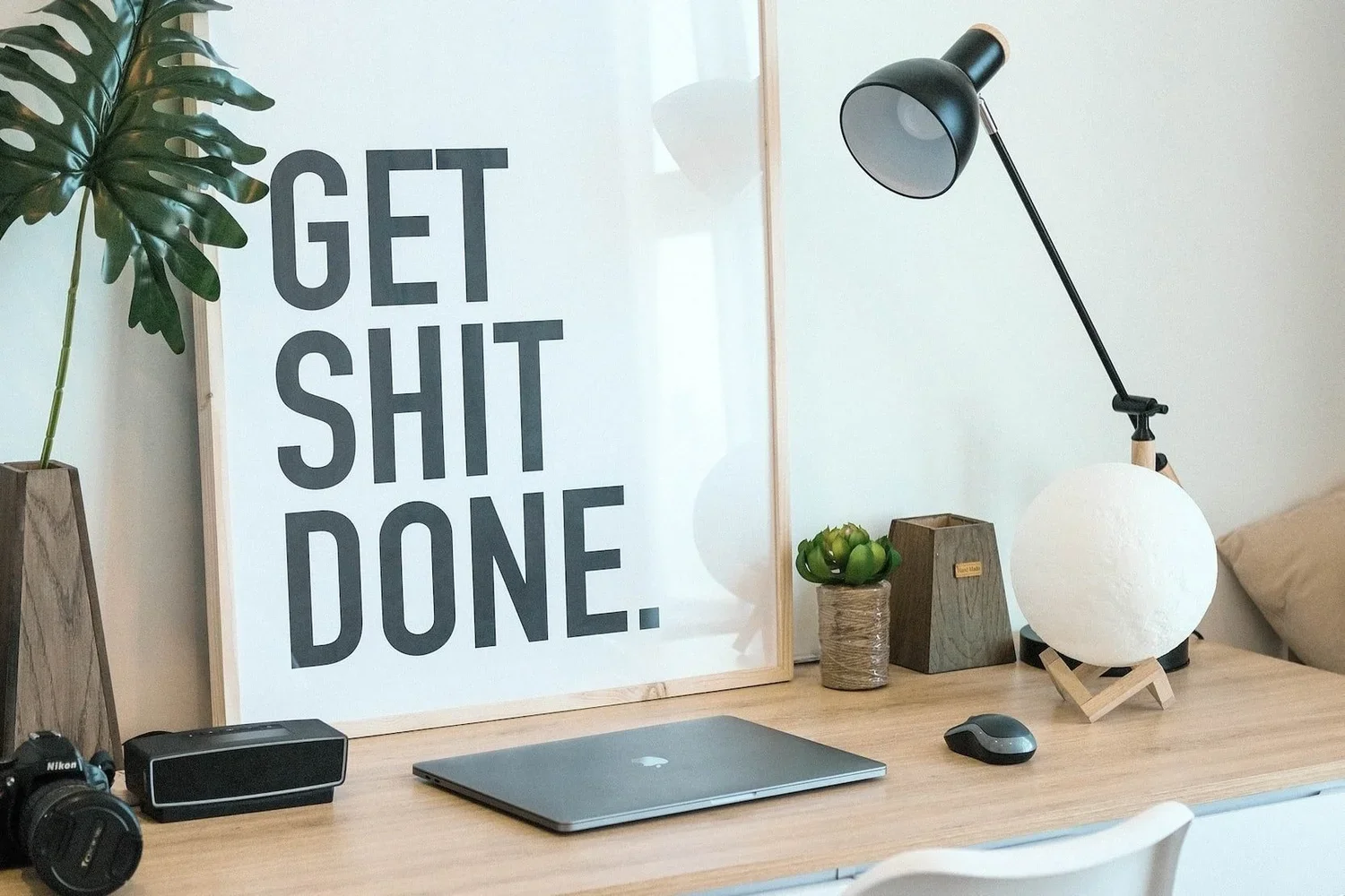 A modern home office desk with a large framed wall art that reads 'GET SHIT DONE.' A black desk lamp, a small potted plant, a wooden box, and a decorative spherical lamp are on the desk. There is also a closed laptop, a wireless mouse, and a camera on the desk.