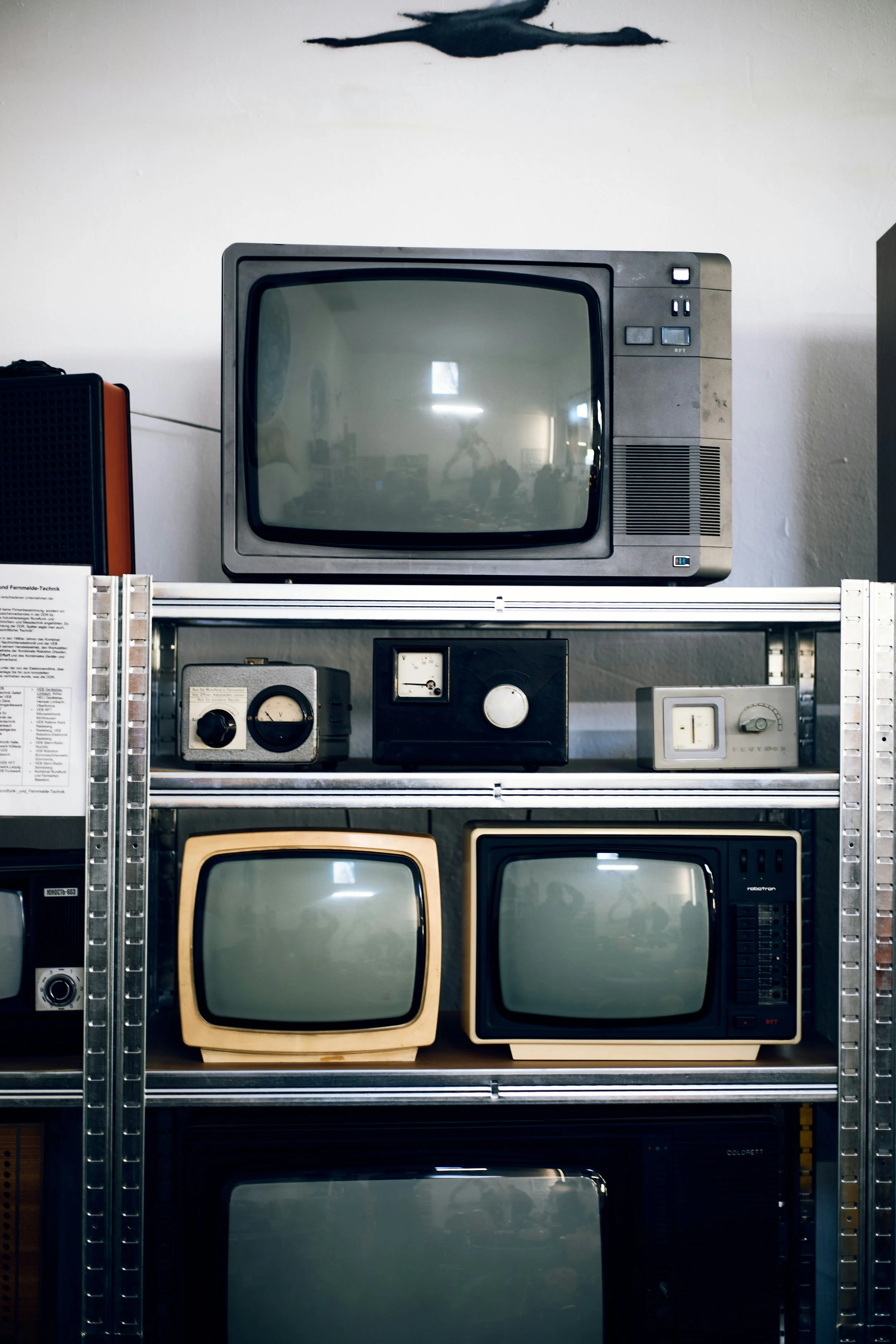 Collection of vintage televisions and electronic devices on metal shelves, with ceiling fan above in background.