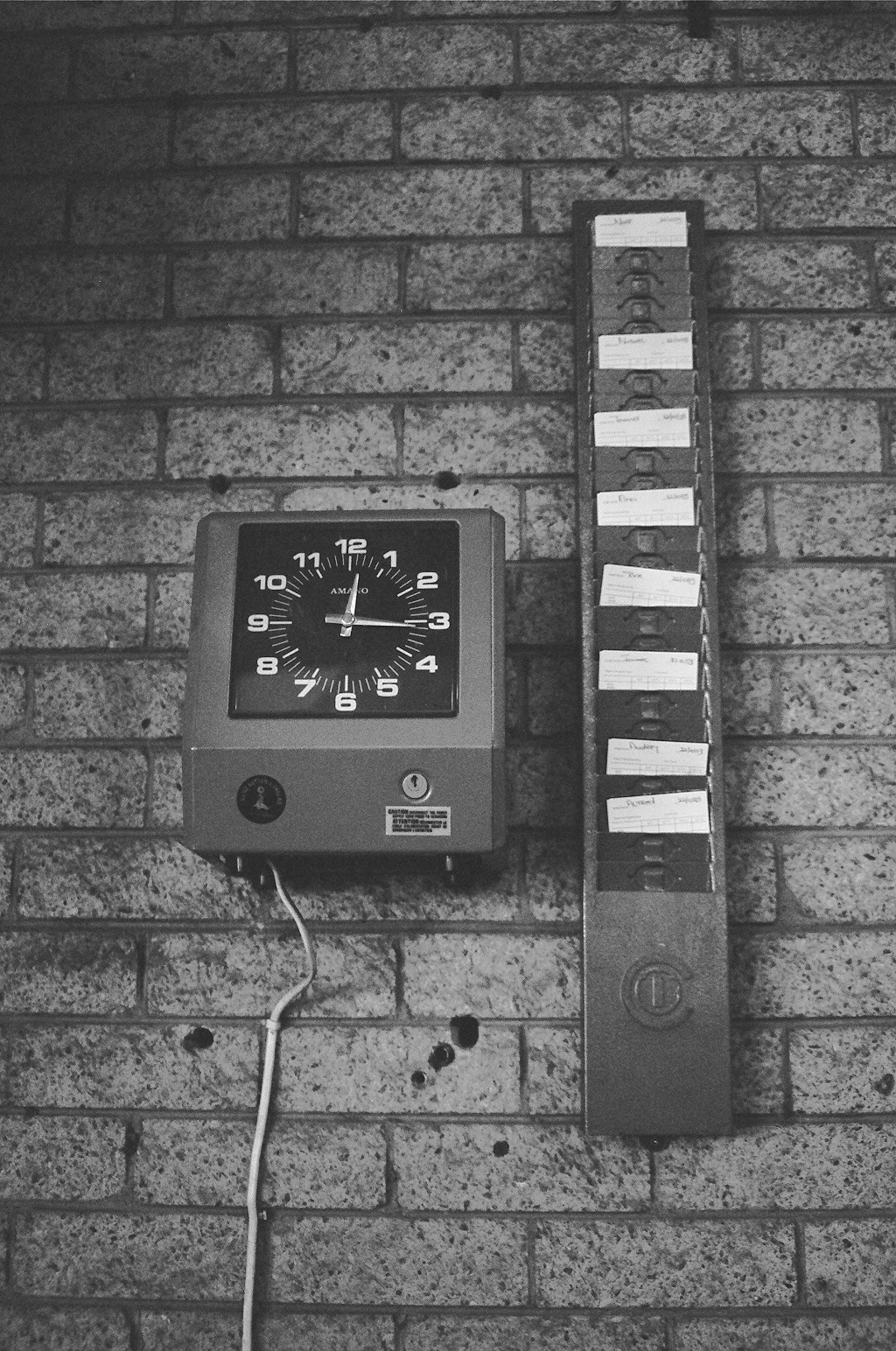 A black and white photo of an old-style time clock with a timecard rack on a brick wall
