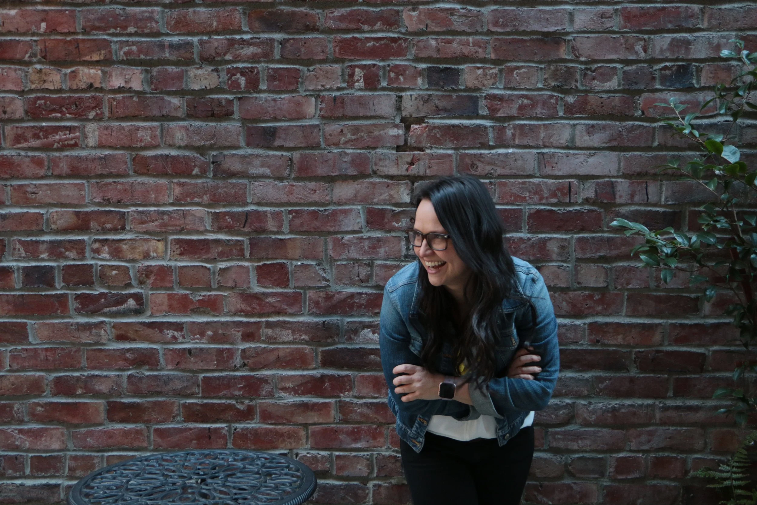 Rachel Bearbower of Nonprofit Automation Agency stands wearing glasses, long dark hair and a denim jacket in front of a brick wall, smiling and laughing.