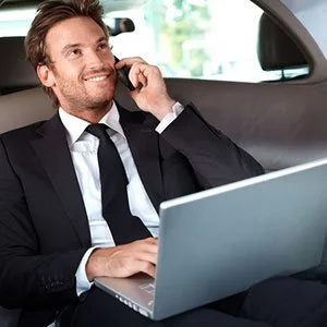 Businessman in a suit sitting in the backseat of a car, talking on a mobile phone, with a laptop on his lap.