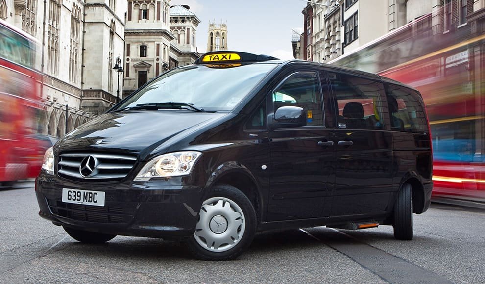 Black Mercedes-Benz taxi van with a yellow taxi sign on top, driving through a city street with blurred red double-decker buses.