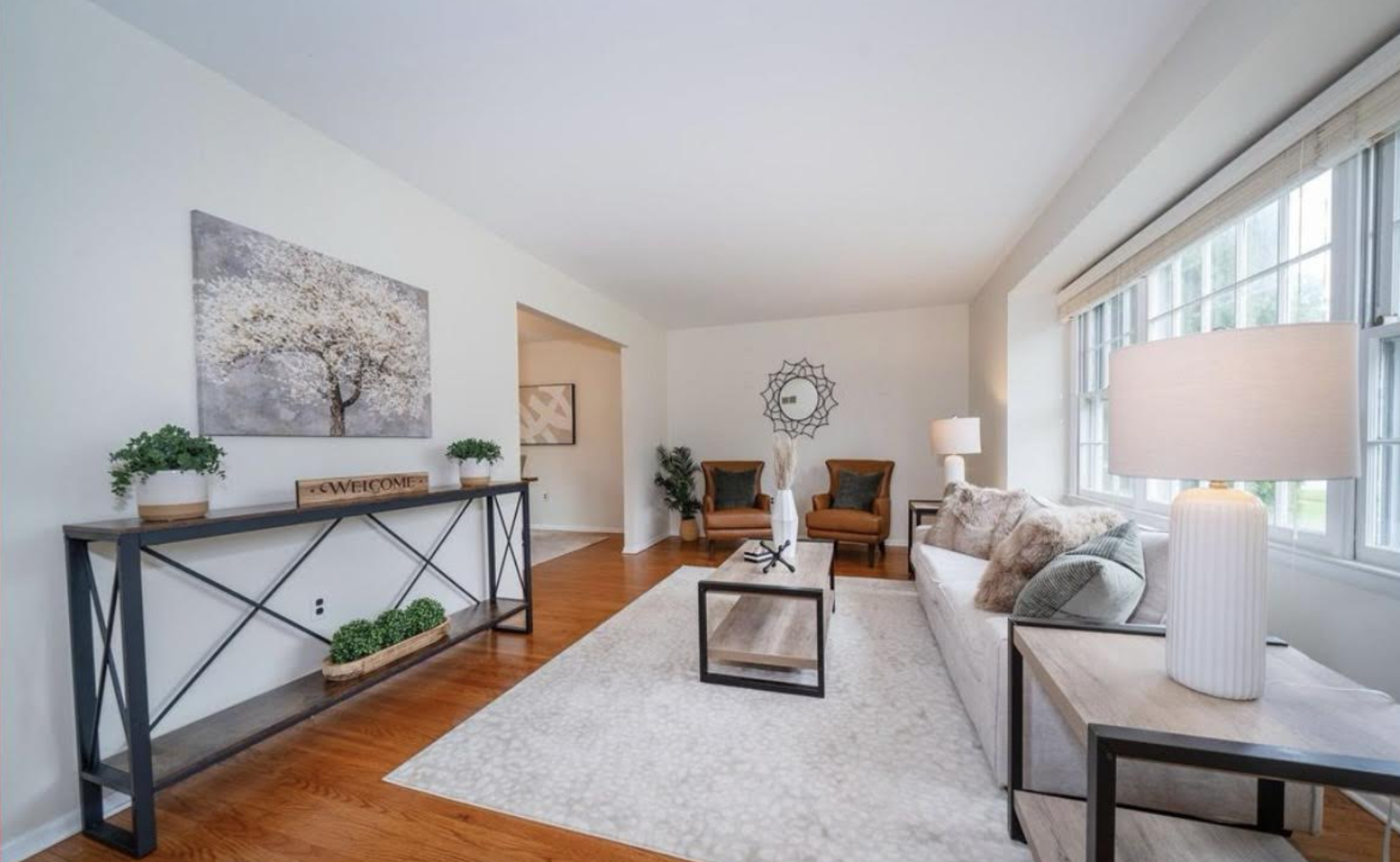 Bright living room with large windows, beige sofa with pillows, two orange armchairs, wooden coffee table, white rug, and side tables, with decorative plants and artwork on walls.