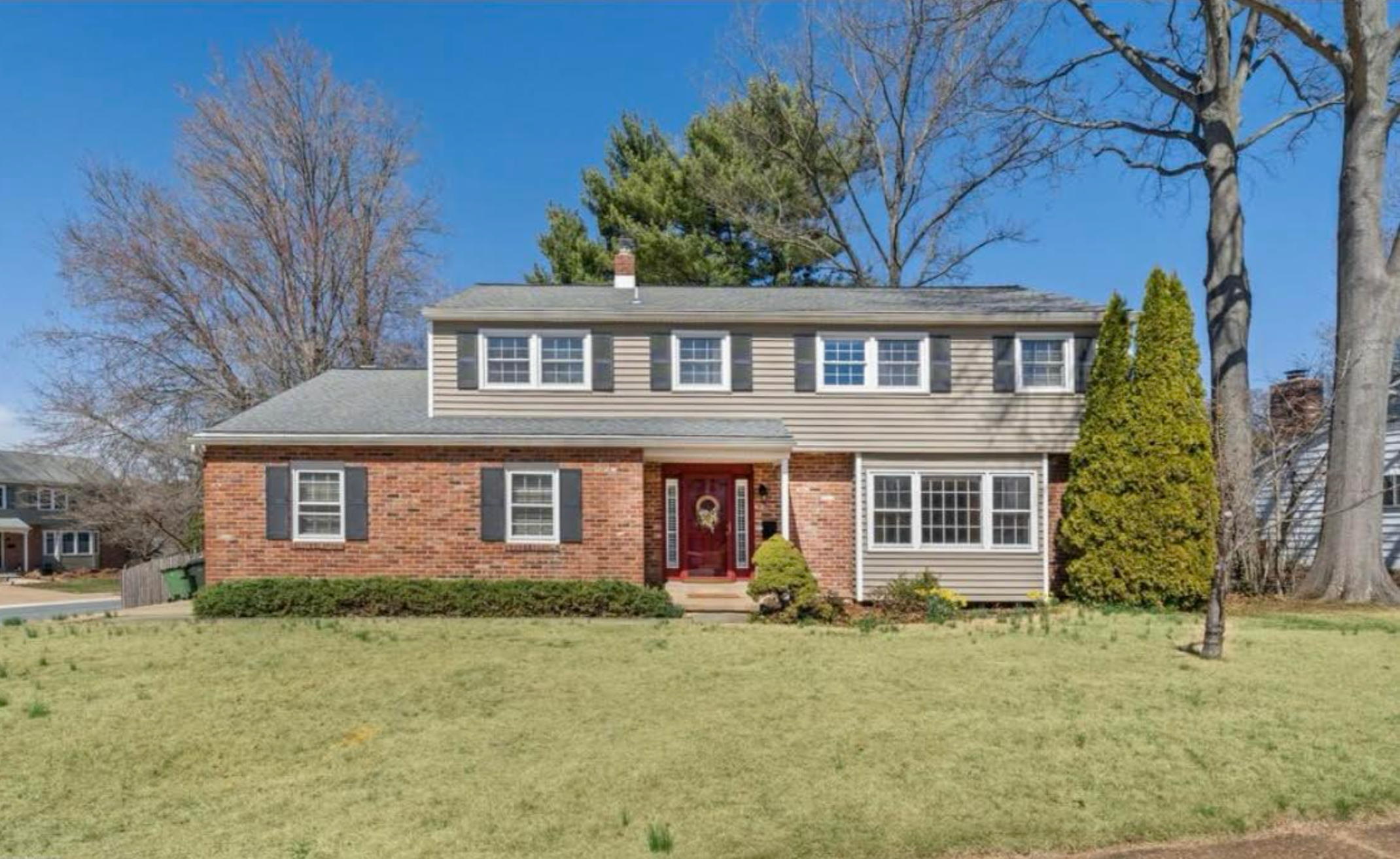 A two-story house with a brick facade on the lower level and beige siding on the upper level, multiple windows with white frames, a red front door with a wreath, and a well-maintained lawn with trees in the background.