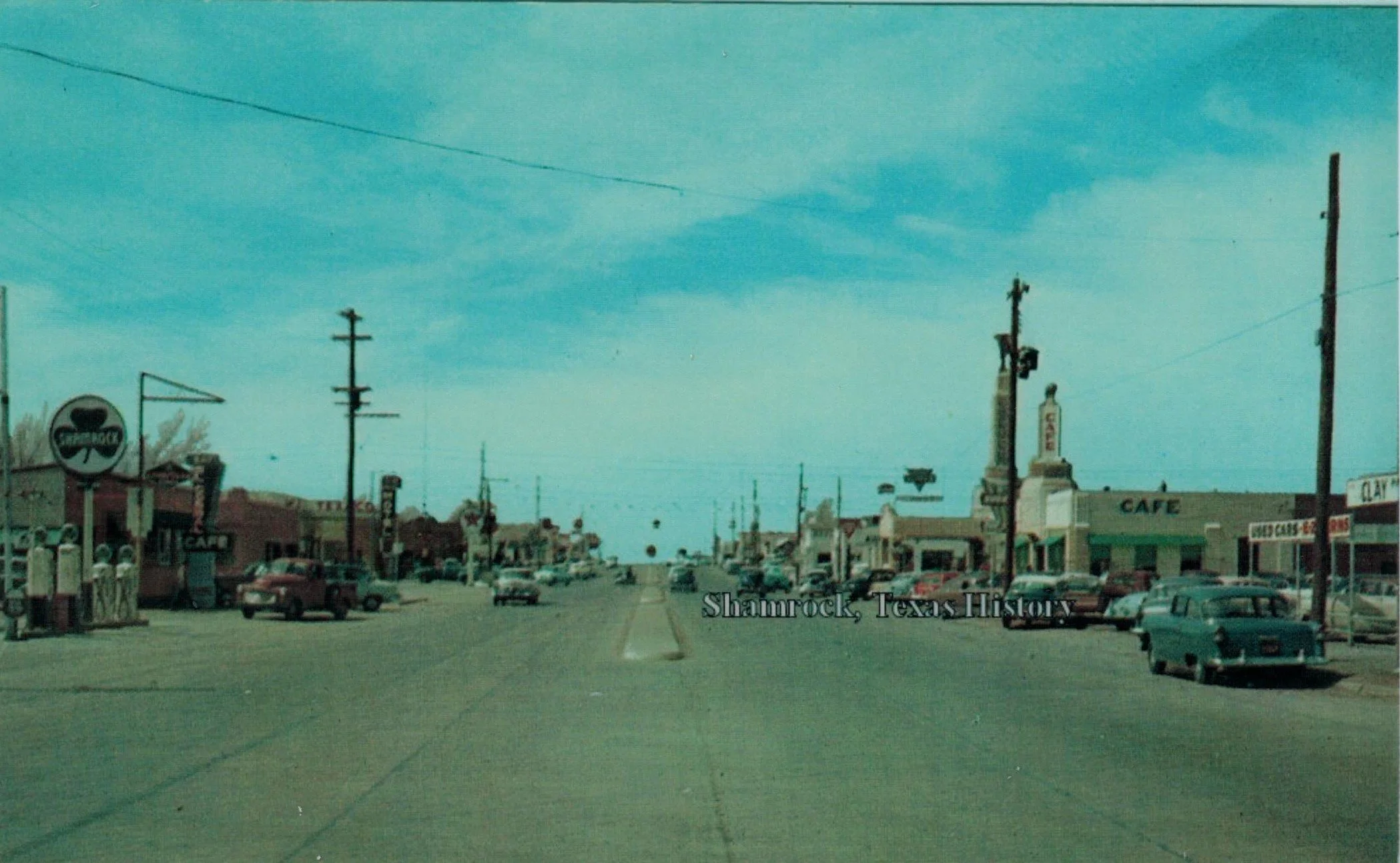 Historic street scene in Shamrock, Texas with vintage cars parked along the road, small businesses, and a clear sky.