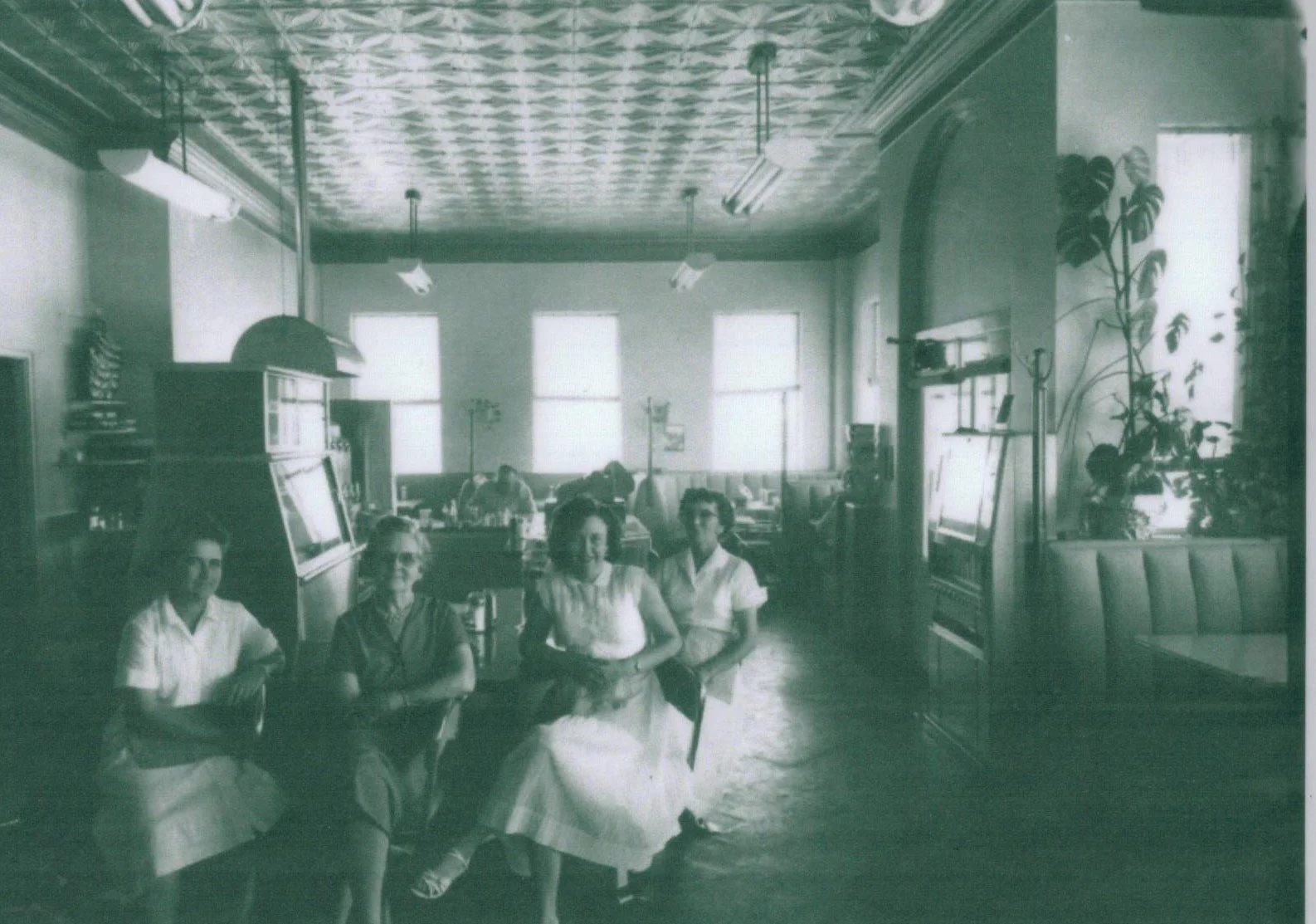 Four women sitting at a table inside a vintage restaurant or cafe, with large windows, patterned ceiling, and vintage furniture and decor.