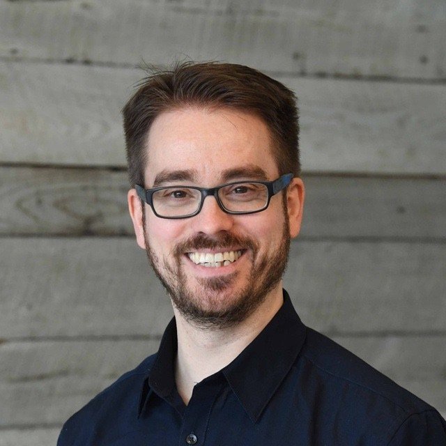 A smiling man with glasses, a beard, short brown hair, wearing a black shirt, standing in front of a wooden wall.