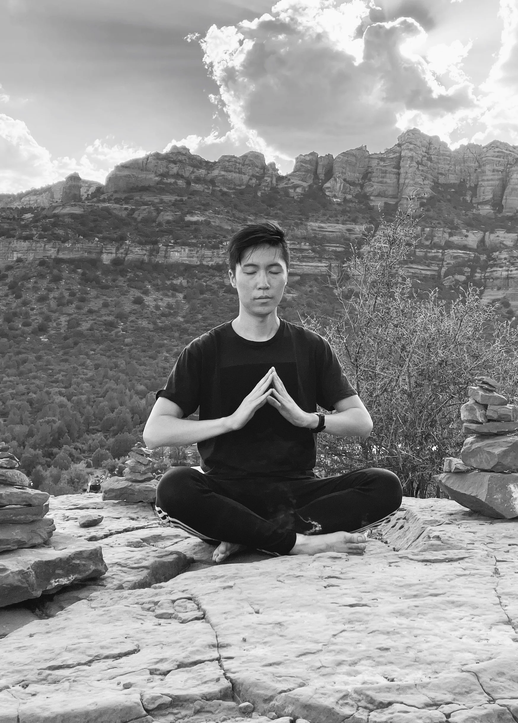 Rayner meditating in a seated cross-legged posture on a rocky surface, with mountains and clouds in the background, outdoors.
