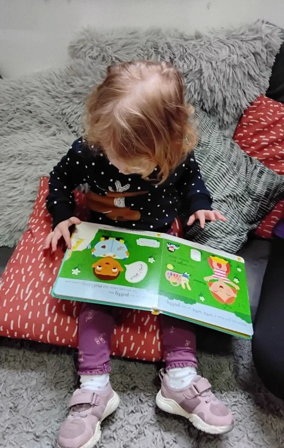 A young girl sitting on a cushion with a gray furry blanket behind her, reading a colourful children's book.