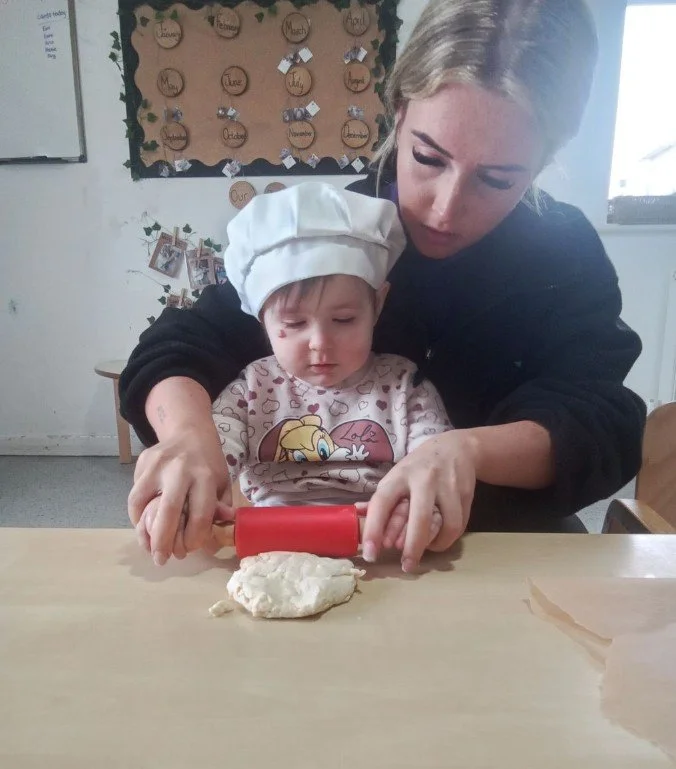 A young child rolling dough out, assisted by a nursery practitioner during a baking activity