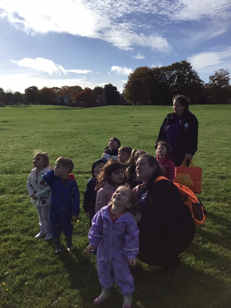 Nursery children exploring outdoors and looking up at the sky with staff during a park outing