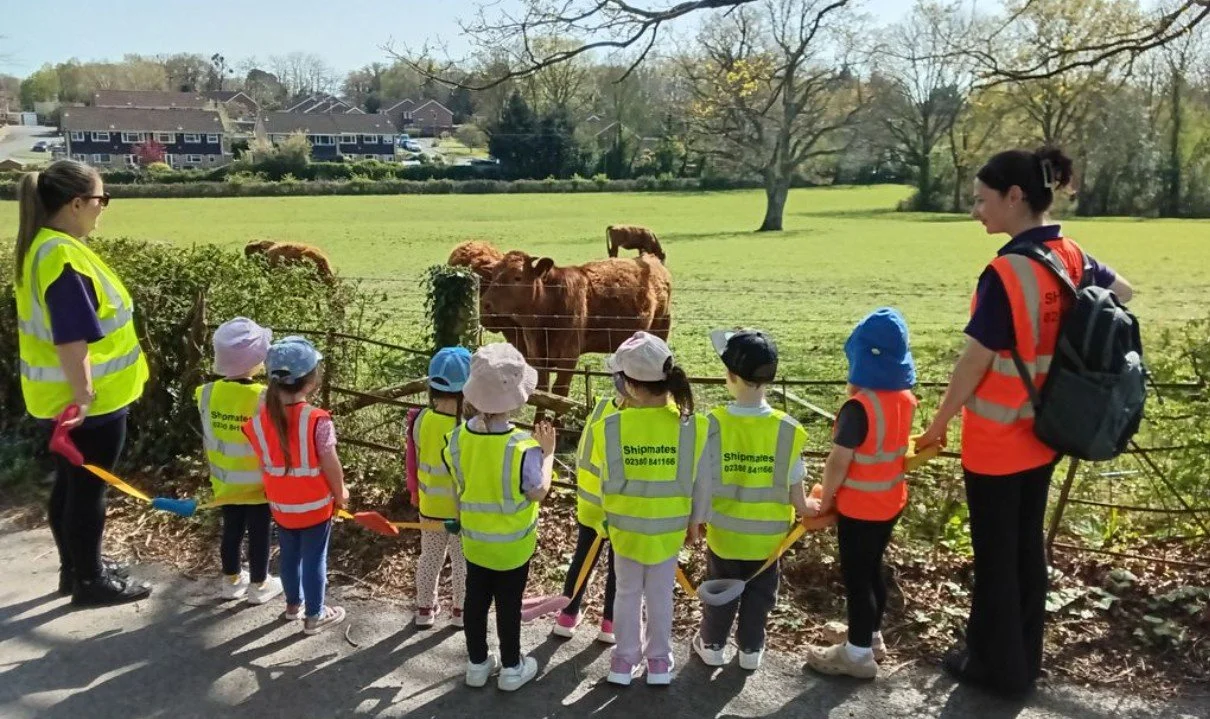 A group of pre-schoolers watching a field of cattle during an excursion, supervised by two nursery practitioners