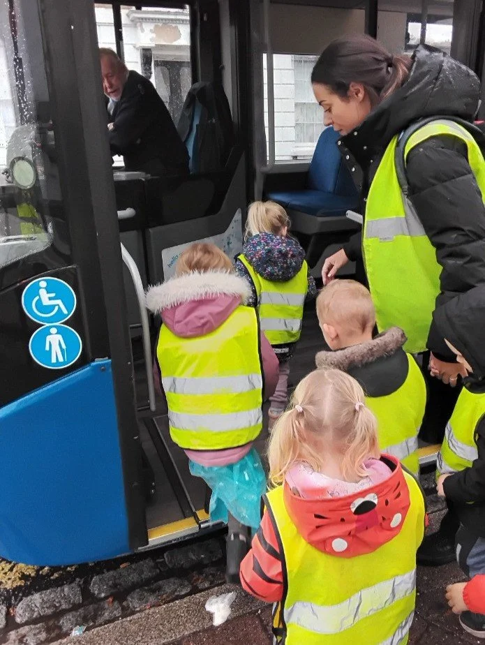 Children wearing yellow safety vests boarding a bus, guided by a nursery practitioner during a fun excursion.