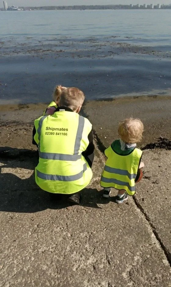 Nursery practitioner crouching with two children by the water’s edge during a supervised outing, all wearing high-visibility jackets