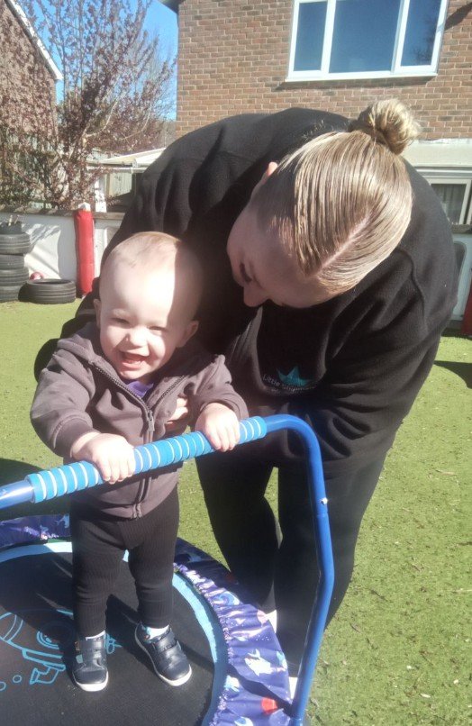 A smiling baby girl holding onto a blue handlebar on a small trampoline, with a nursery practitioner close by, outdoors in the garden