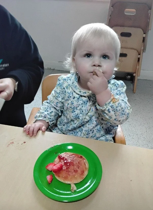 A young girl sitting in a chair at a table, eating from a plate of food during snack