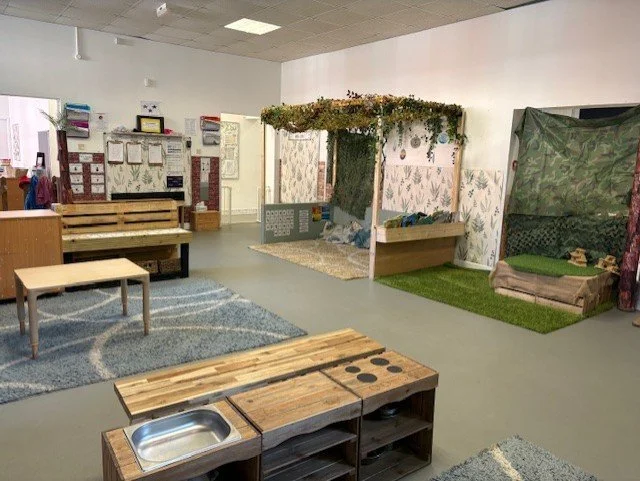 Indoor classroom with a pretend play area resembling a small house with a canopy and foliage decoration, wooden benches, a play kitchen, and area rugs.