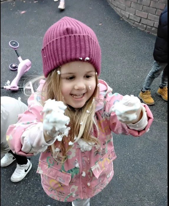 A young girl smiling and laughing as she plays with foam during outdoor play.