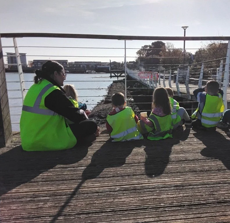 Children from day nursery sitting on a pier with a practitioner during a supervised outing, wearing high-visibility jackets and enjoying refreshments