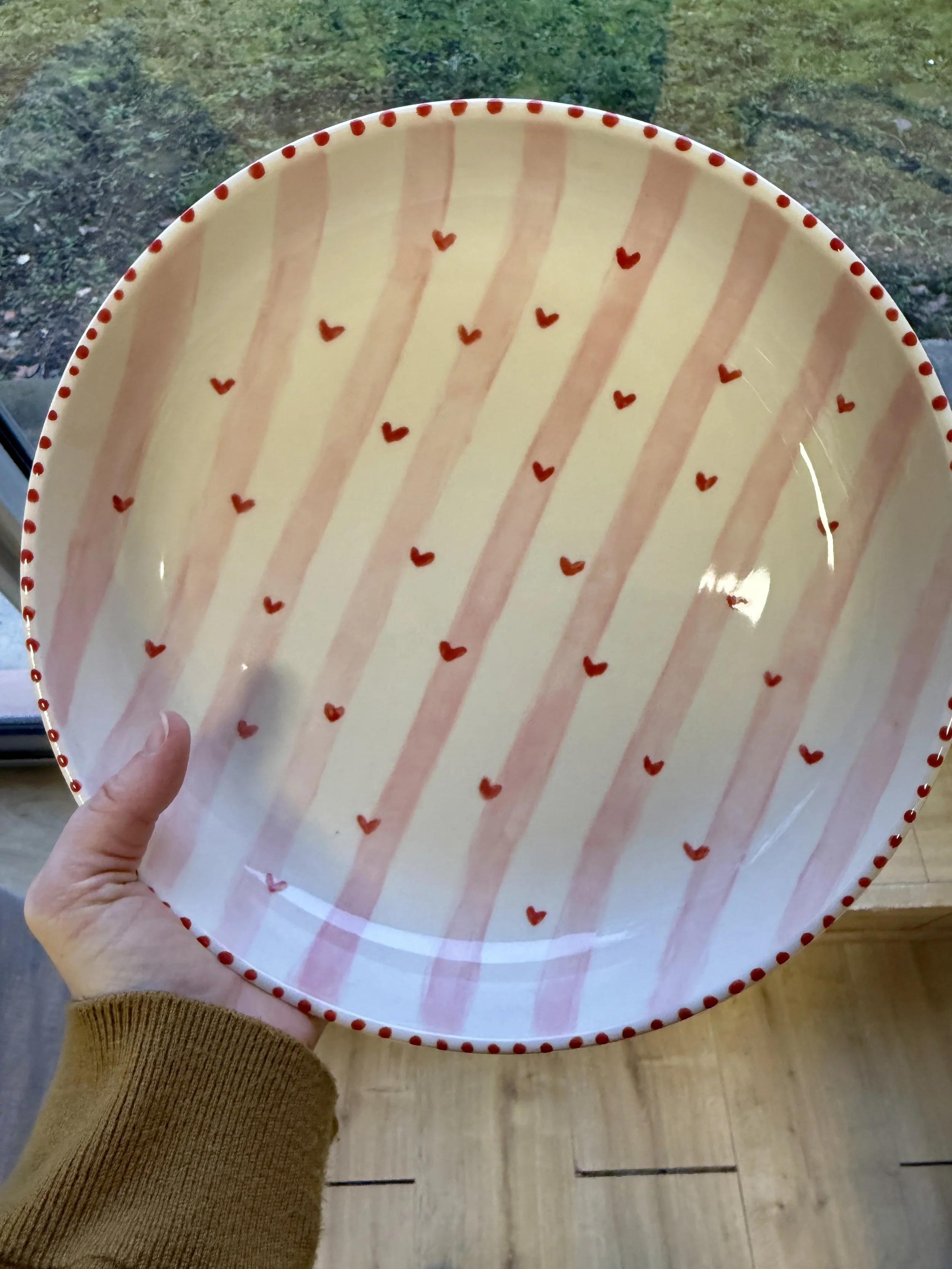 Une assiette décorée de rayures roses et de petits cœurs rouges, tenue par une main avec un pull beige, devant une surface en bois et une fenêtre avec vue sur l'extérieur.