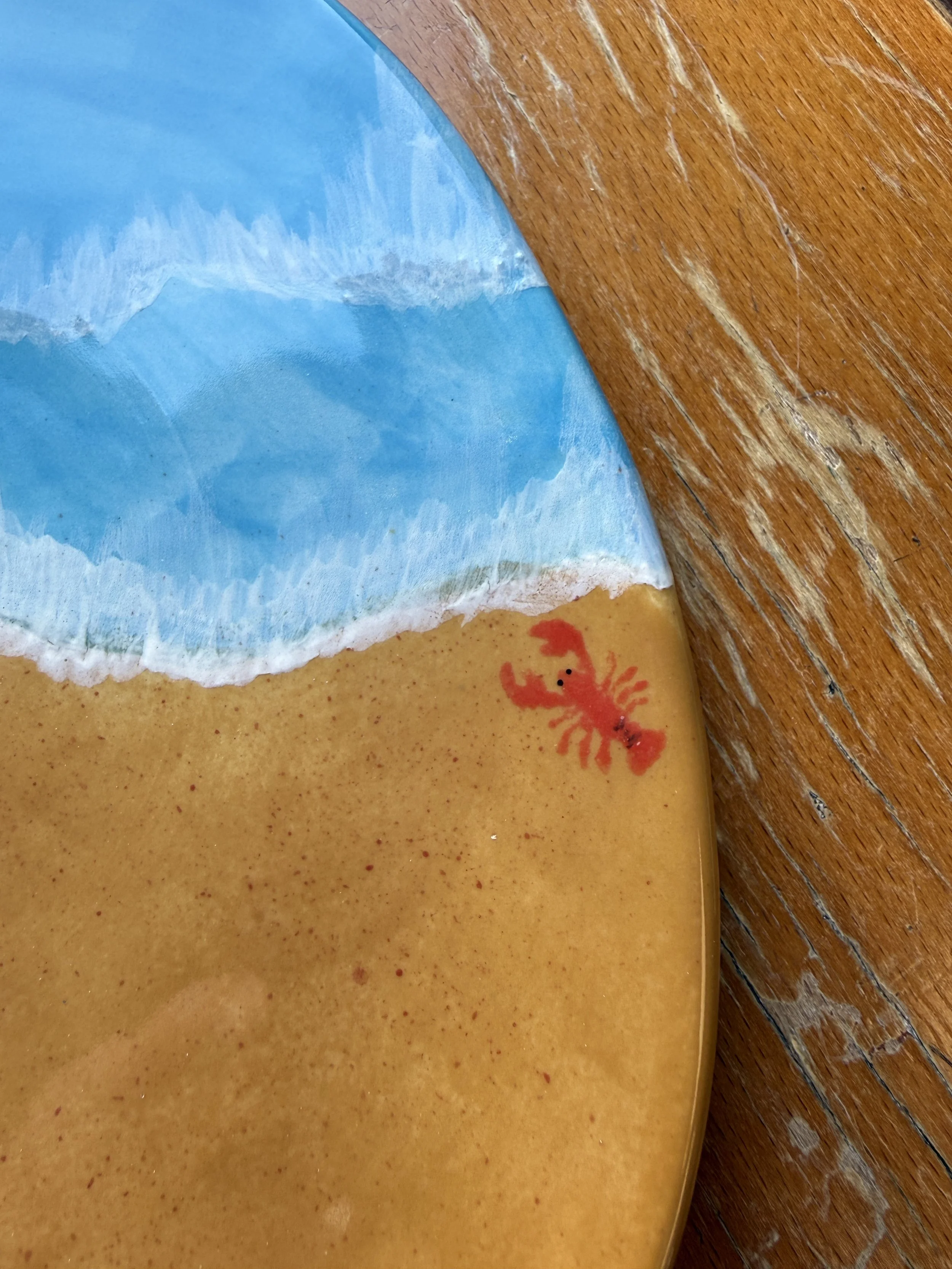Assiette en céramique peinte avec des vagues bleues et du sable, avec un dessin de langoustine rouge, posé sur une table en bois.