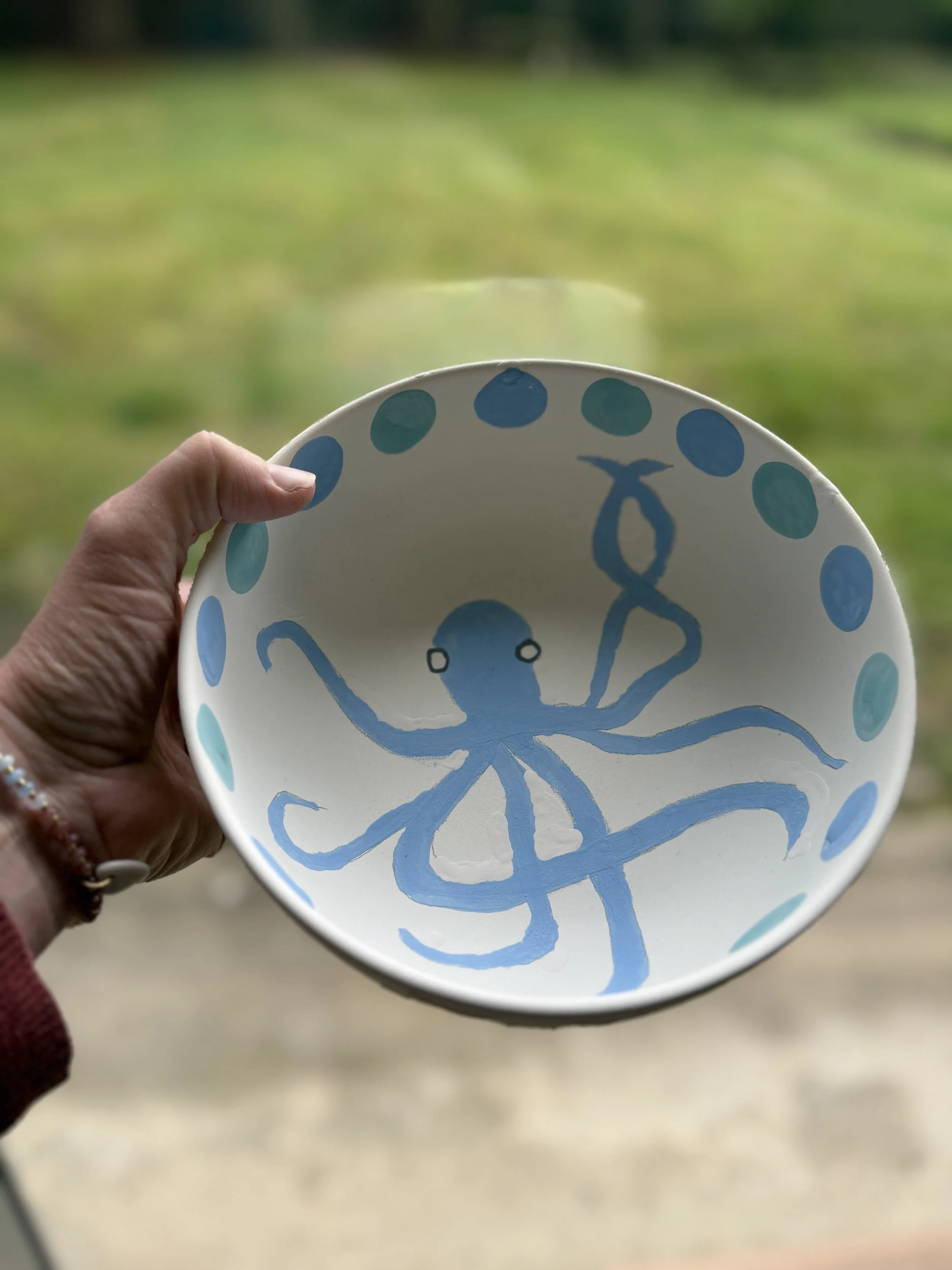 Assiette en céramique blanche avec dessin d'un poulpe en bleu et pois bleus autour, tenue par une main devant un fond flou de verdure.