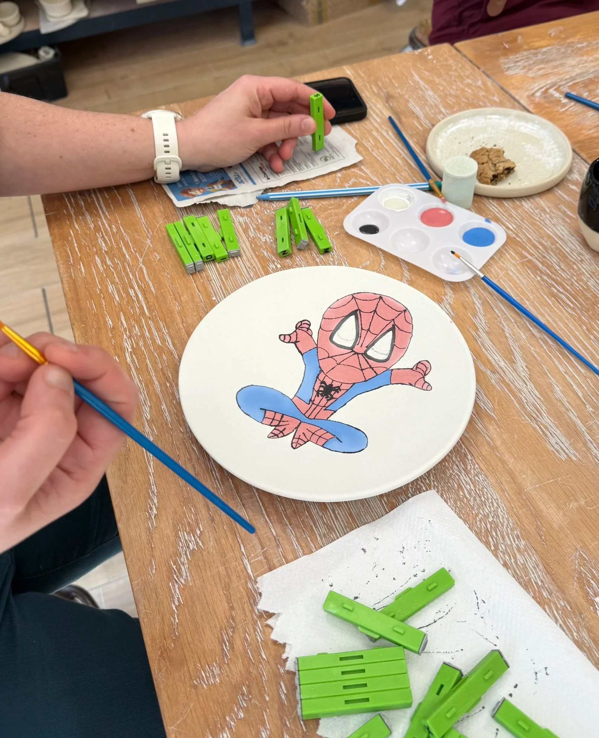 Un atelier artistique avec une personne peignant une assiette avec un dessin de Spider-Man, entourée de pinceaux, des blocs de peinture verts, une palette de pigments, et divers outils sur la table en bois.