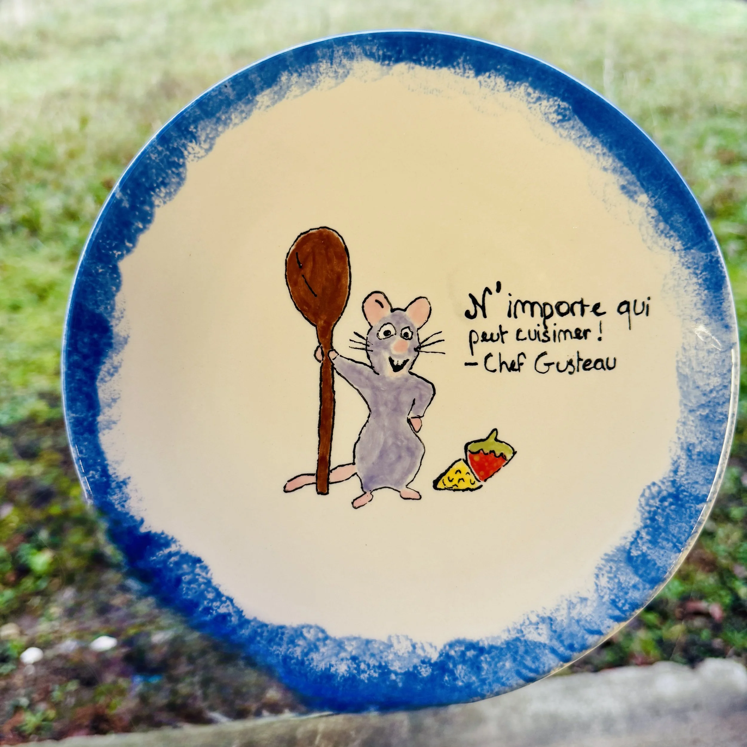 Une assiette avec un dessin d'une souris tenue un cuillère en bois, avec une phrase en français : "N'importe qui peut cuisiner! - Chef Gustau" et quelques aliments à côté.