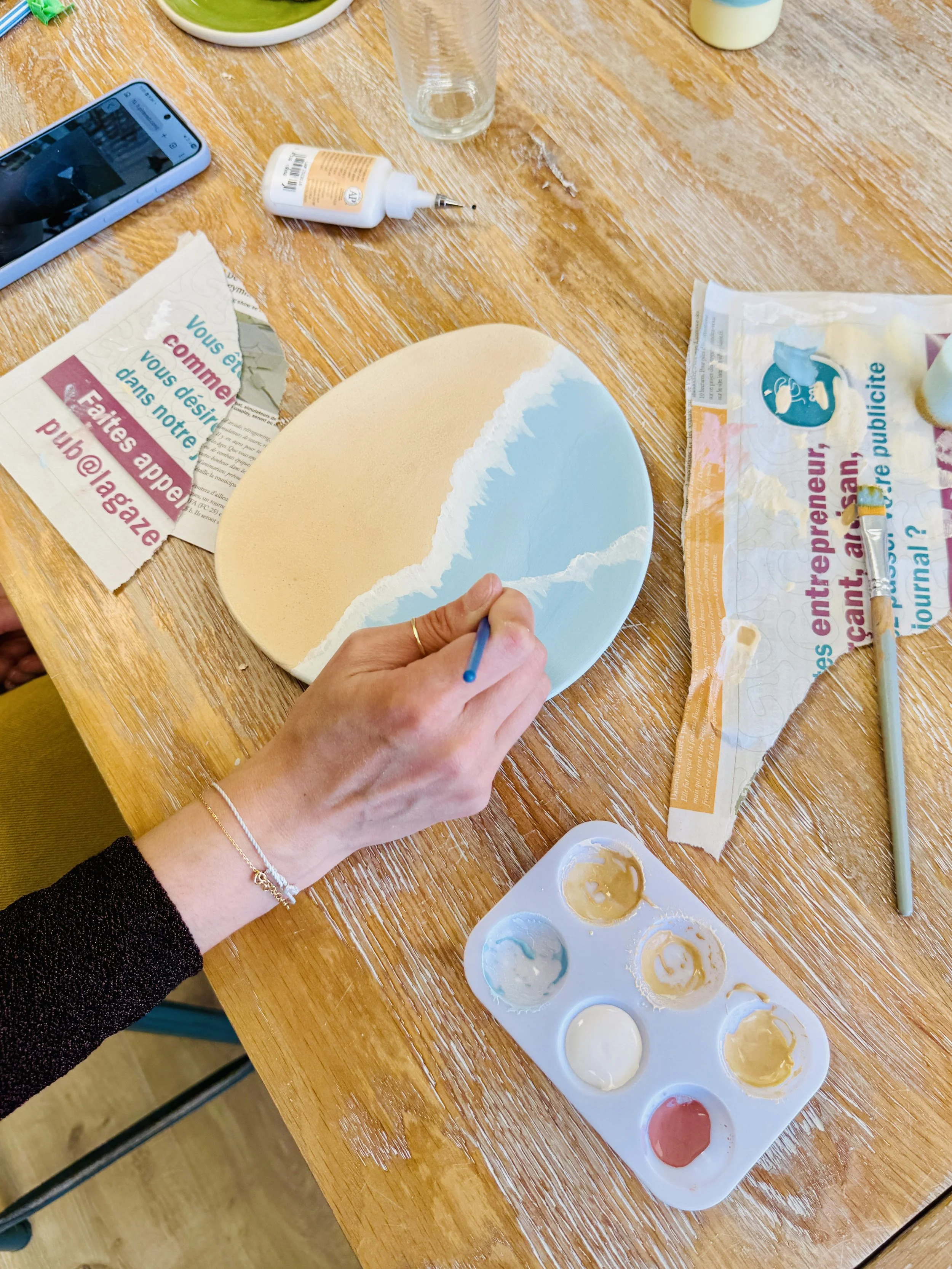 Une personne peint une assiette ronde en utilisant des couleurs pastel, avec une palette de peinture contenant plusieurs couleurs, sur une table en bois. Autour, il y a des objets divers, comme un smartphone, un tube de colle, un verre vide, du papie