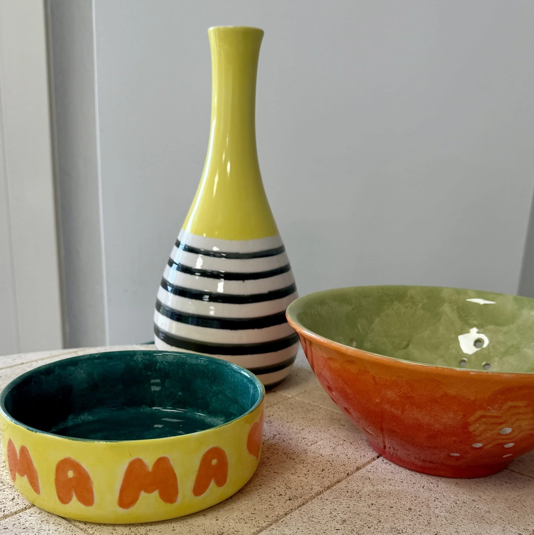 Trois récipients en céramique colorés sur une surface en bois. Une grande coupe bicolore orange et vert, une petite boîte jaune avec des lettres orange, et une bouteille à décor noir et blanc avec un haut jaune.