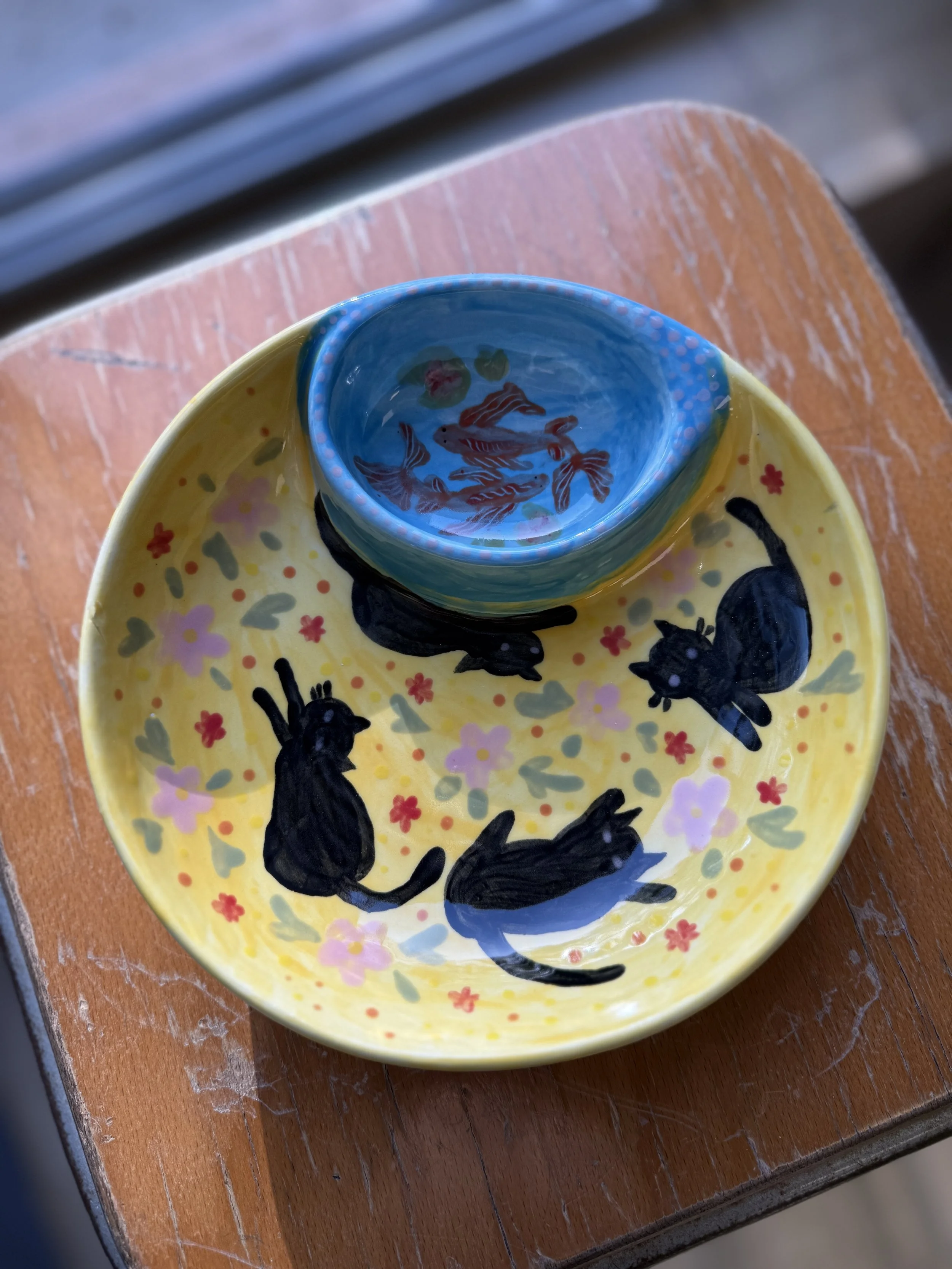 Une assiette jaune décorée de chats noirs et de fleurs, avec un bol bleu contenant de petits poissons rouges, placé sur une table en bois. Il y a une fenêtre en arrière-plan.