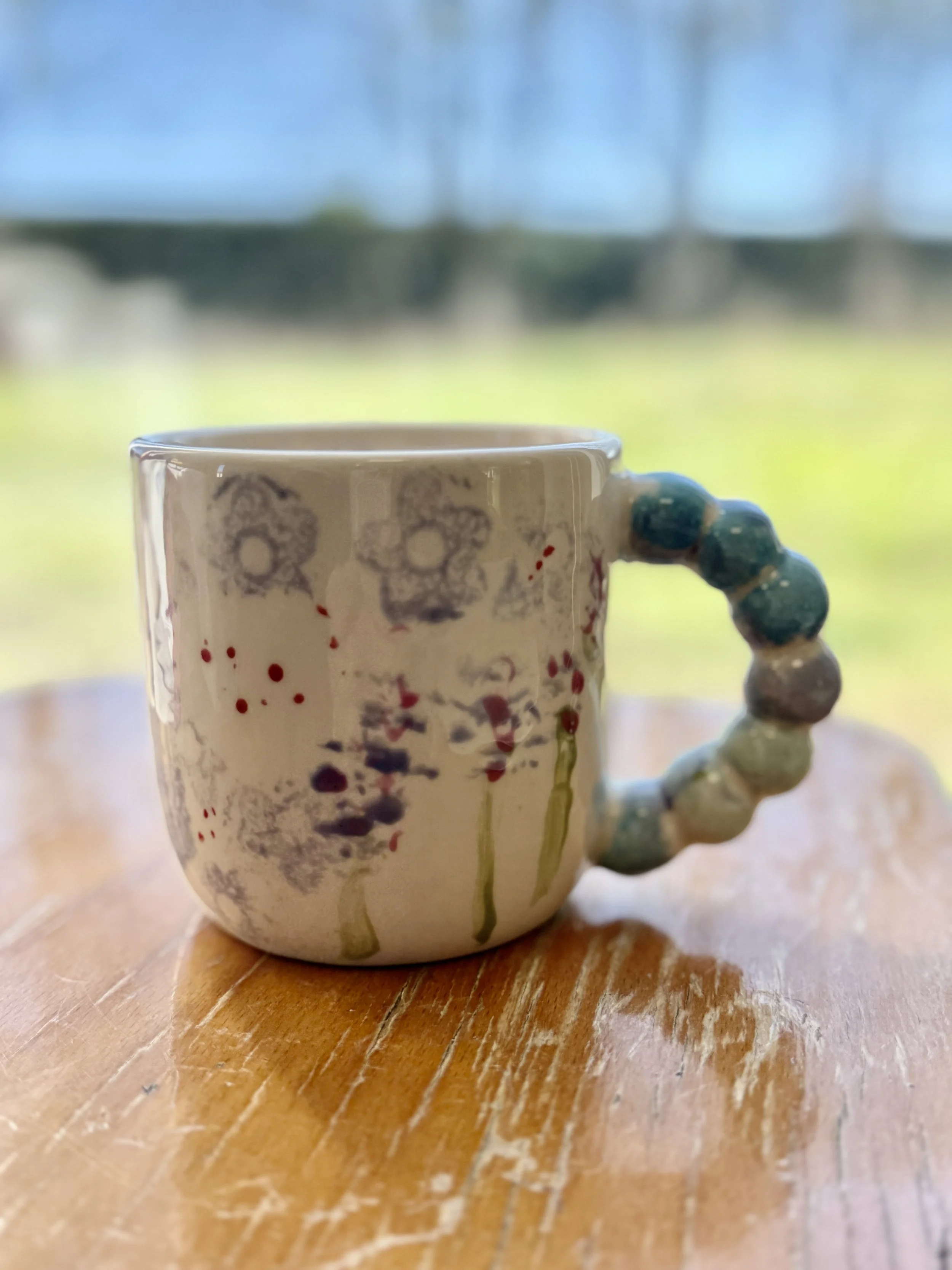 Tasse en céramique blanche avec un motif de fleurs grises, taches rouges et gouttes de peinture vertes, posé sur une table en bois.